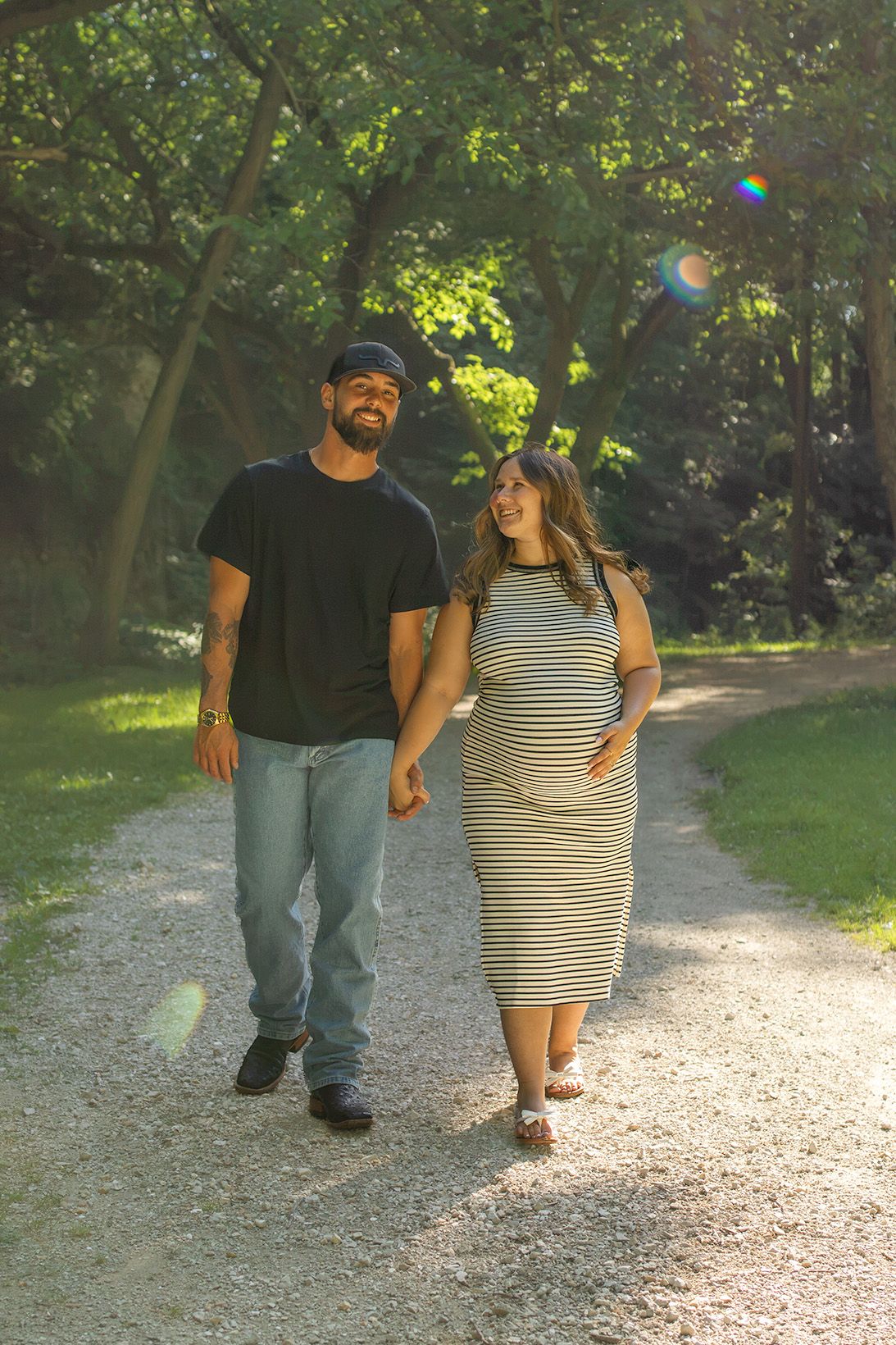 A happy couple walks hand-in-hand on a sunlit park path, the woman visibly pregnant in a striped maxi dress.