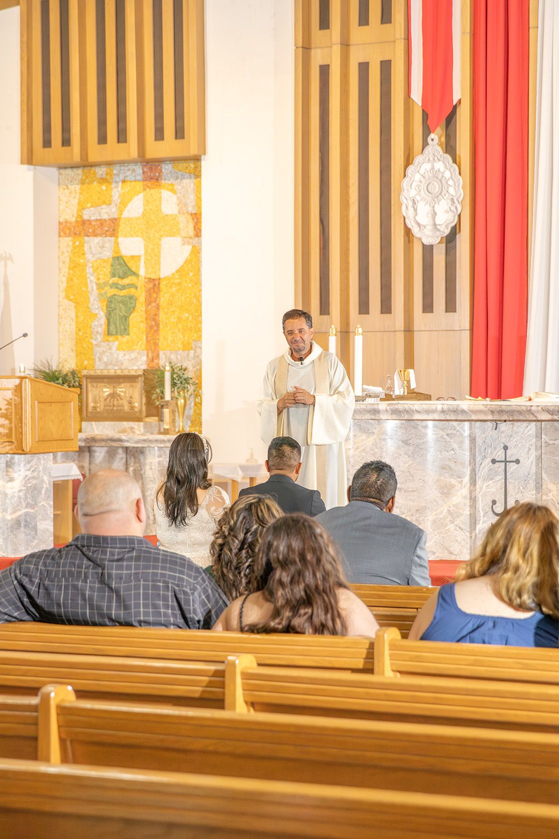Priest in white vestments leads Catholic Mass at altar while congregation sits in wooden pews inside a church.