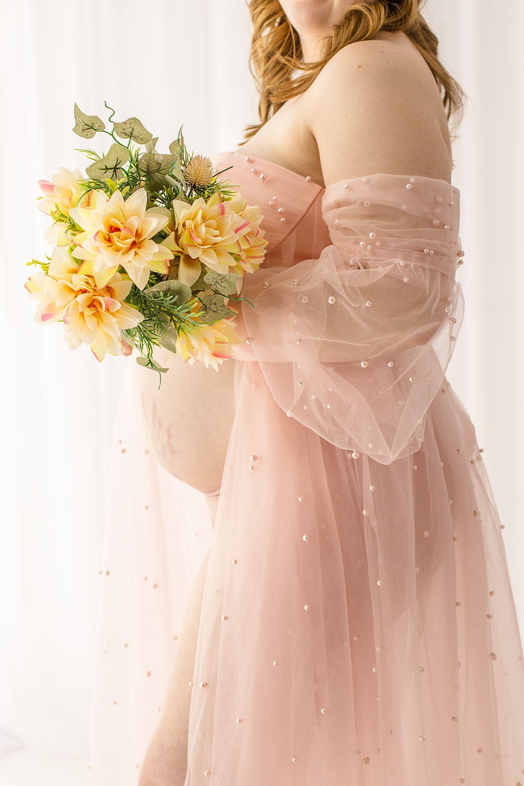 Elegant pink tulle dress with pearl details holds a bouquet of yellow dahlias and greenery against white backdrop.