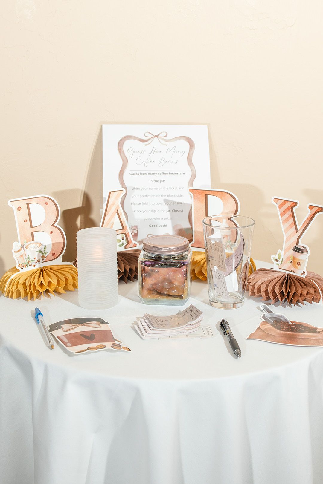Baby shower table with rose gold BABY letters, candles, jars, and activity cards on a white tablecloth.