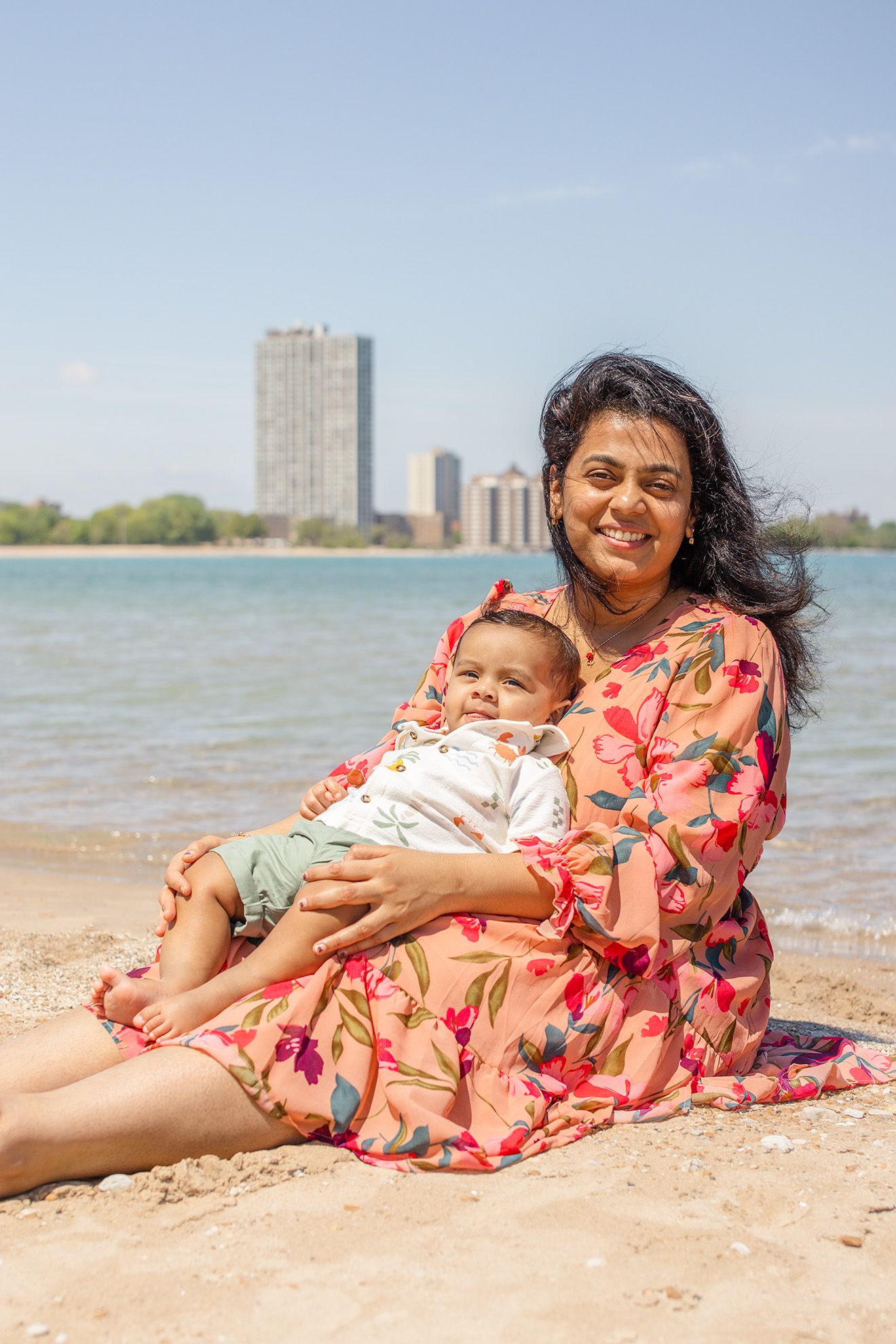 Smiling mother in floral dress sits on sandy beach holding baby, with city skyline and blue water behind.