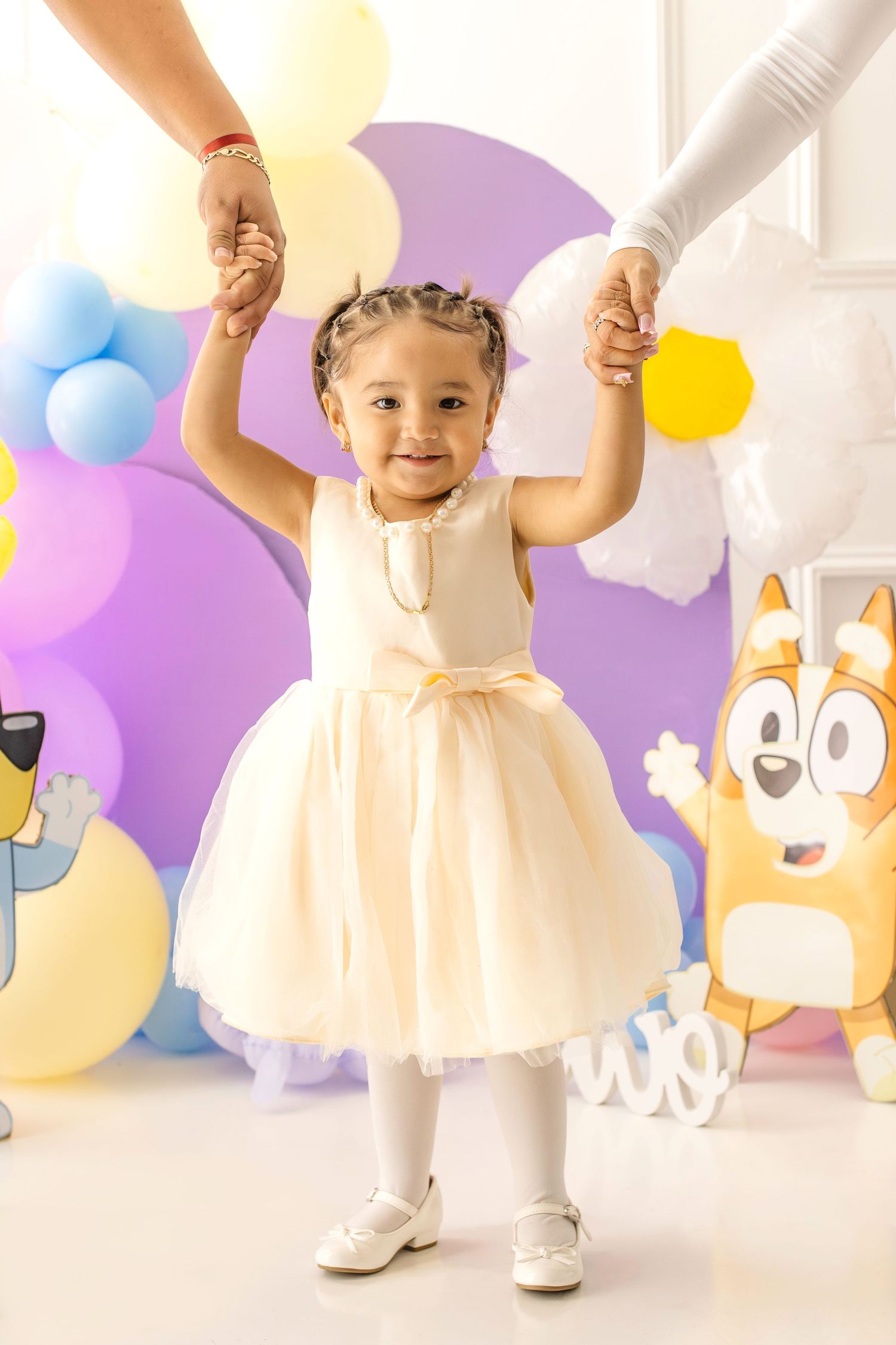 Toddler girl in cream dress holds adults hands at Bluey themed birthday party with colorful balloons.