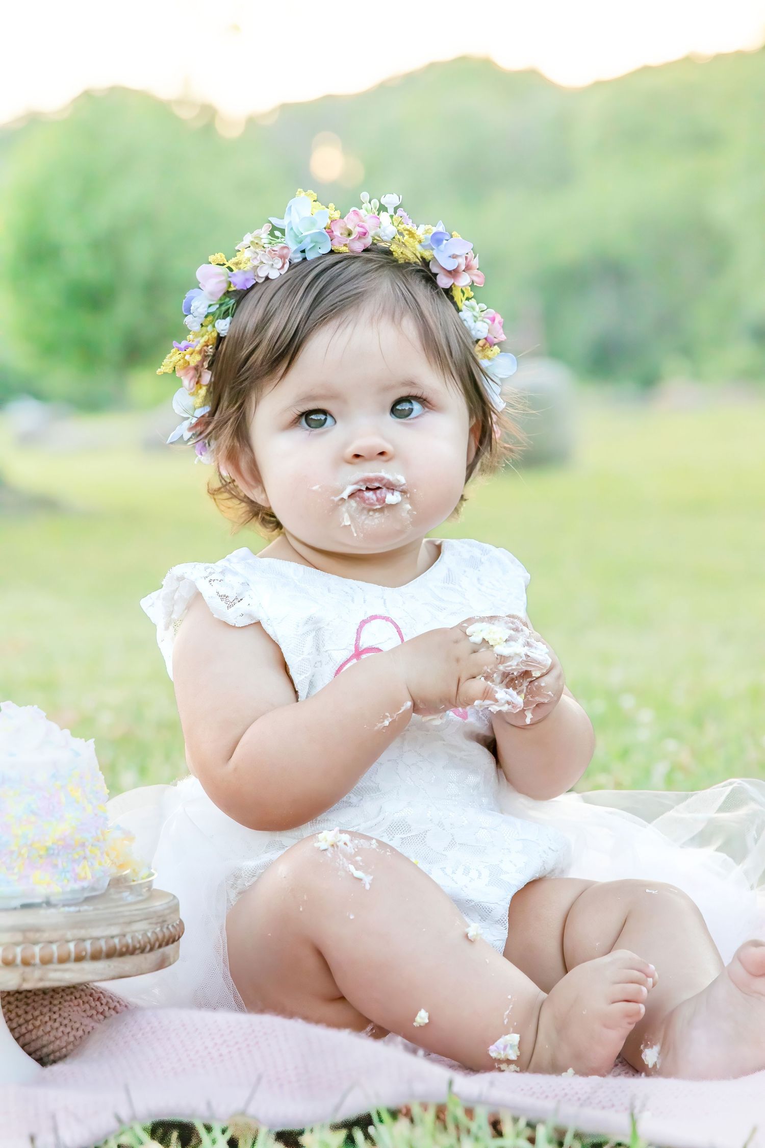 Baby in white dress wearing floral crown sits on pink blanket outdoors with cake smeared on face.