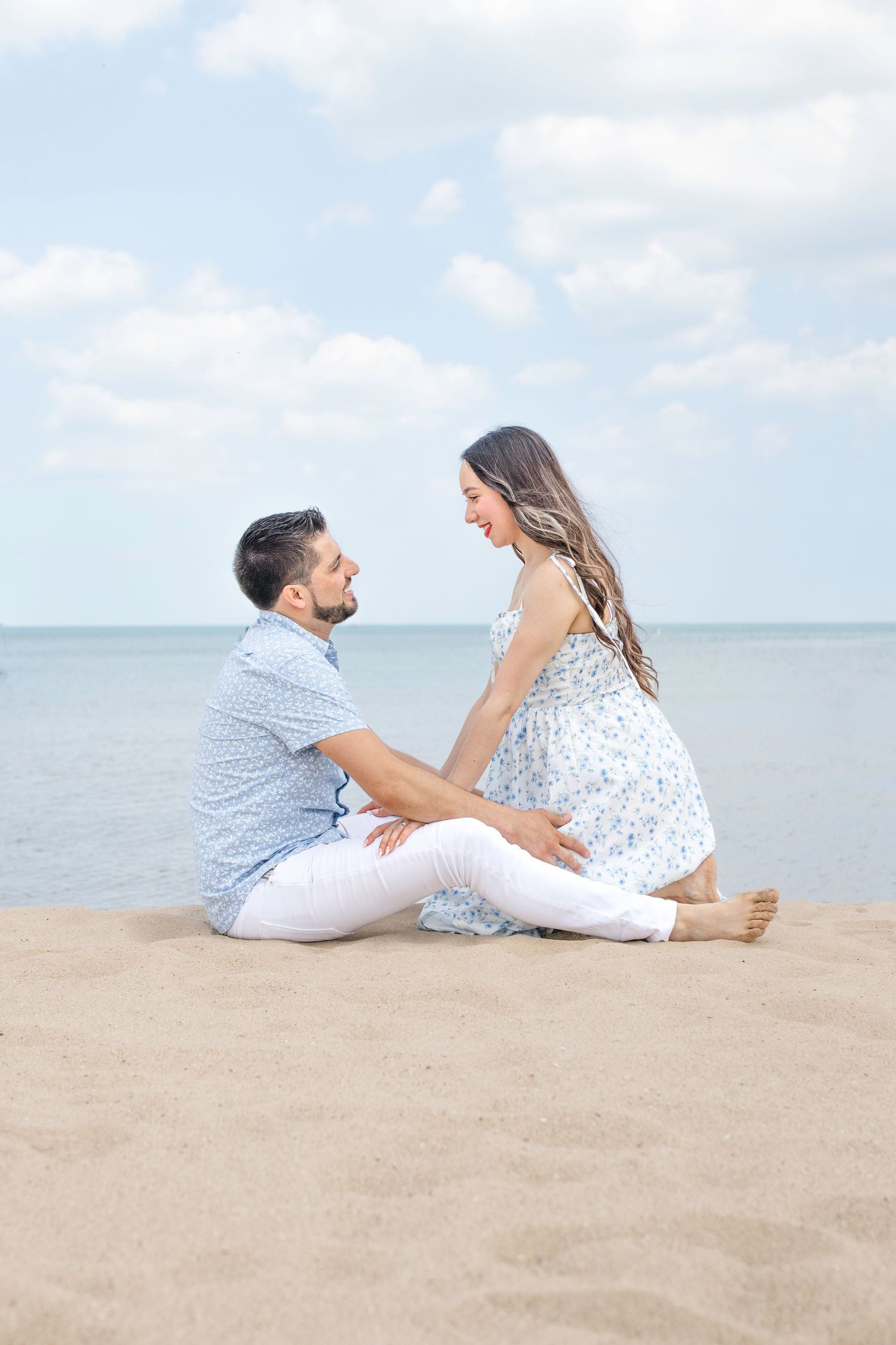 A romantic couple sits together on a sandy beach by the ocean on a beautiful summer day.