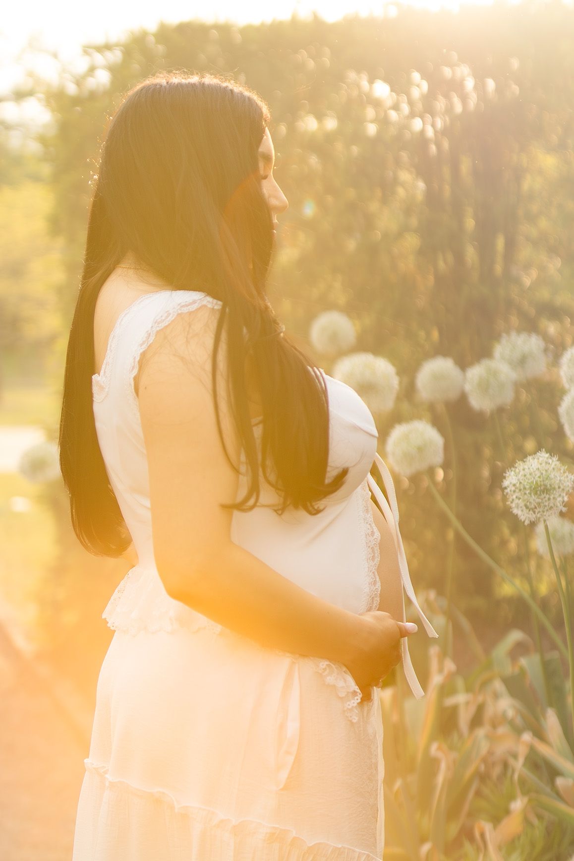 Pregnant woman in white dress holding baby bump in golden sunset light near white allium flowers in garden.