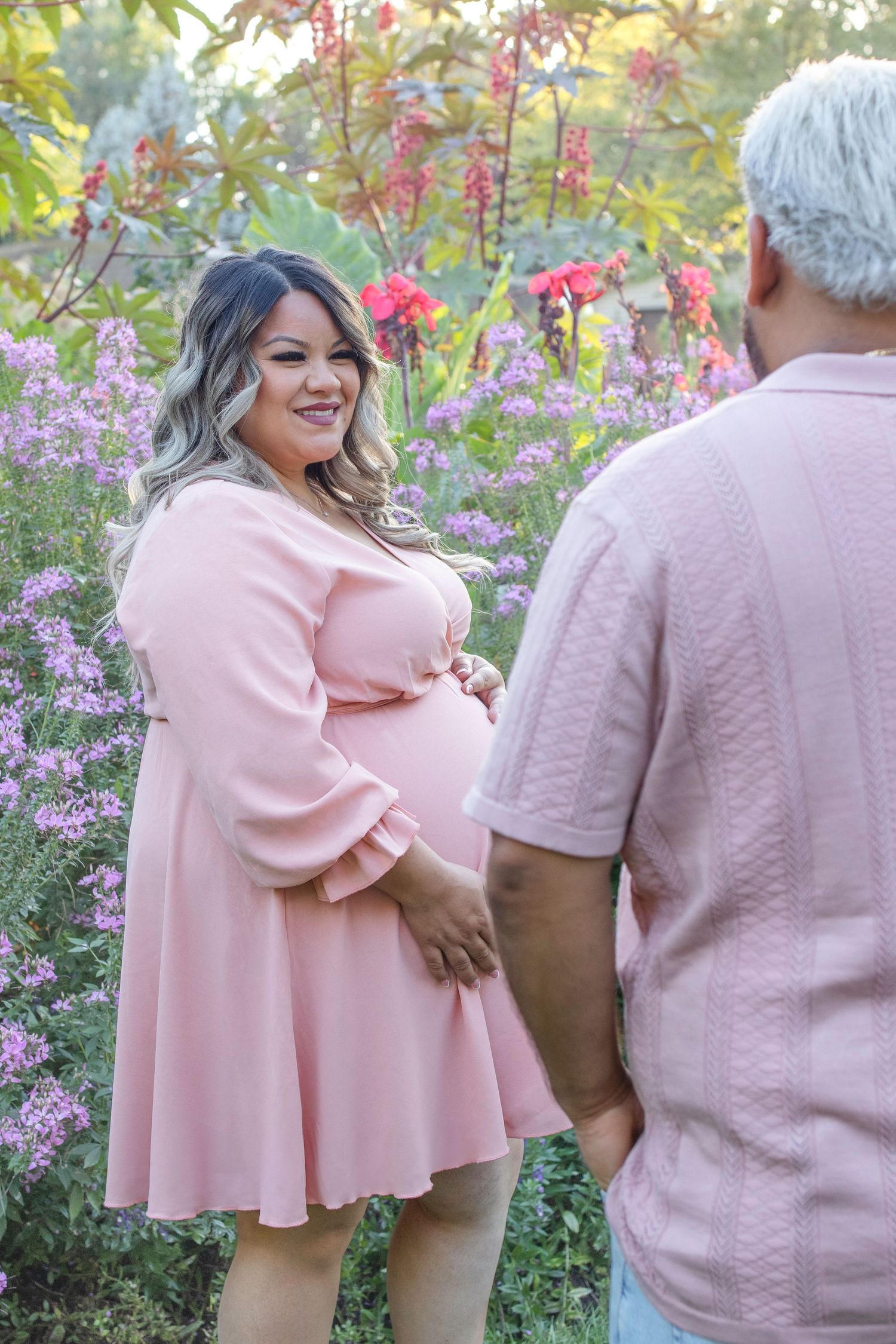 A couple wearing matching pink outfits embraces in a garden with blooming flowers in the background.