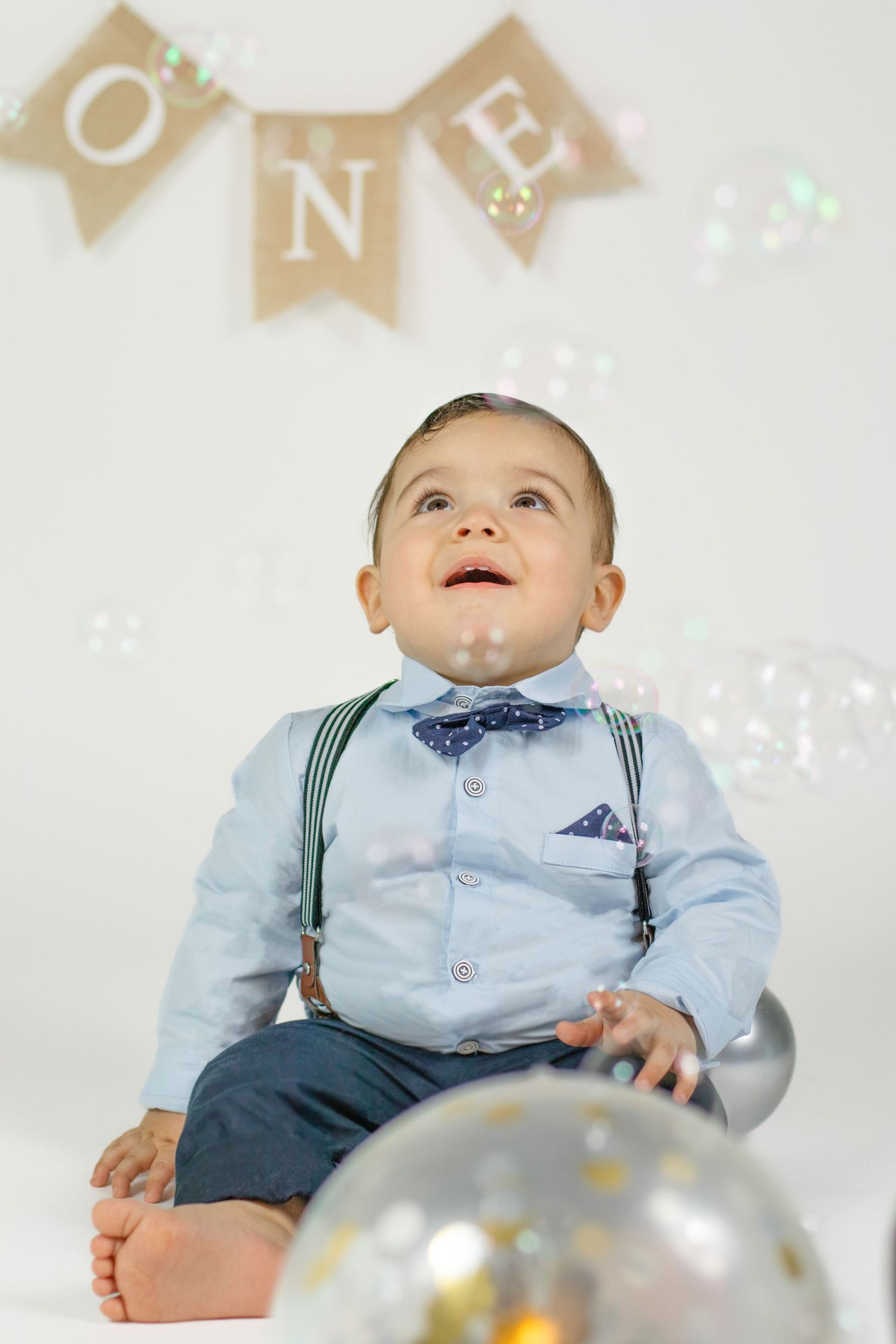 A dressed-up baby boy in suspenders and bow tie celebrates his first birthday with bubbles and a festive ONE banner.