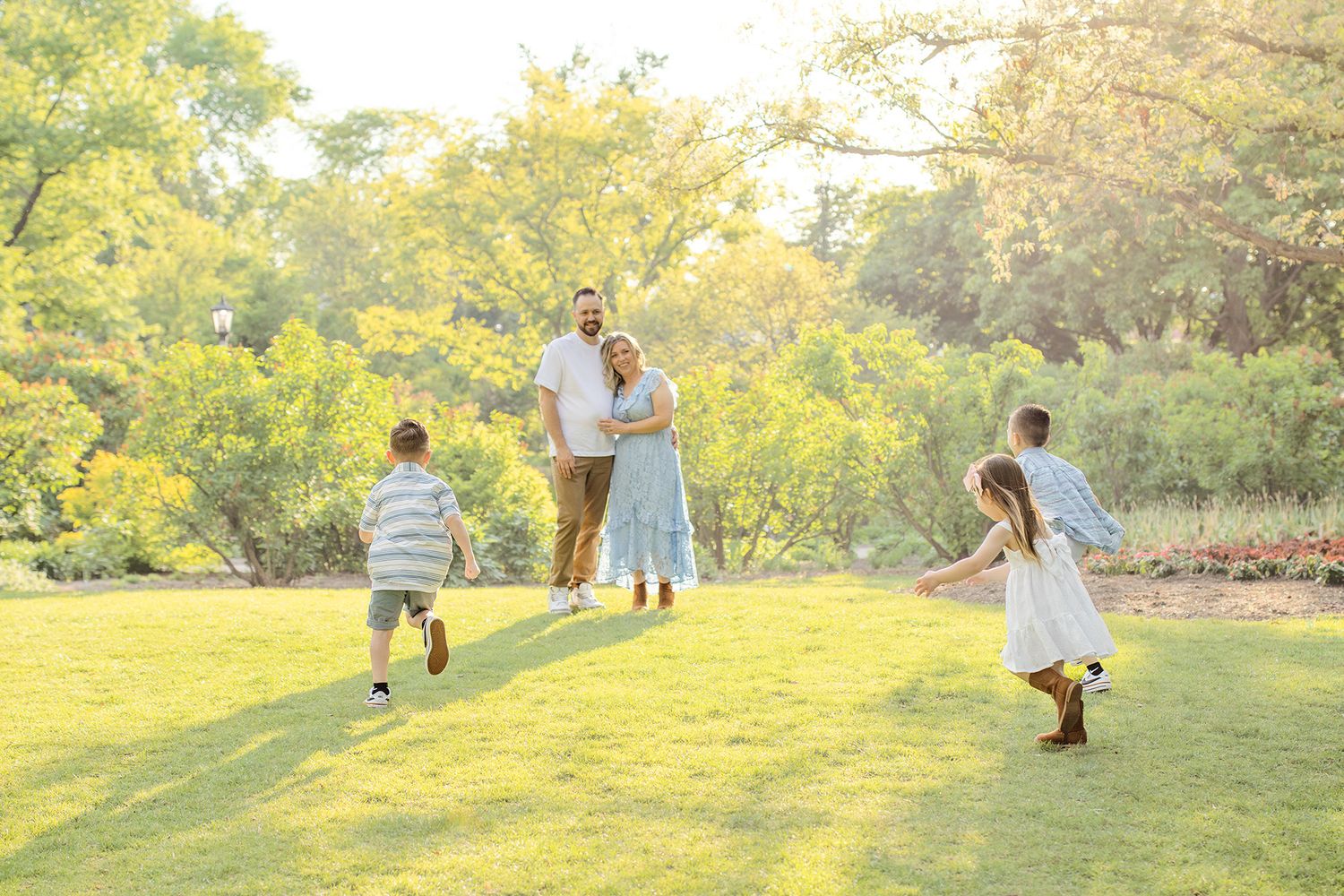 Family enjoying a sunny day outdoors on green grass with two children running toward parents in a lush garden setting.