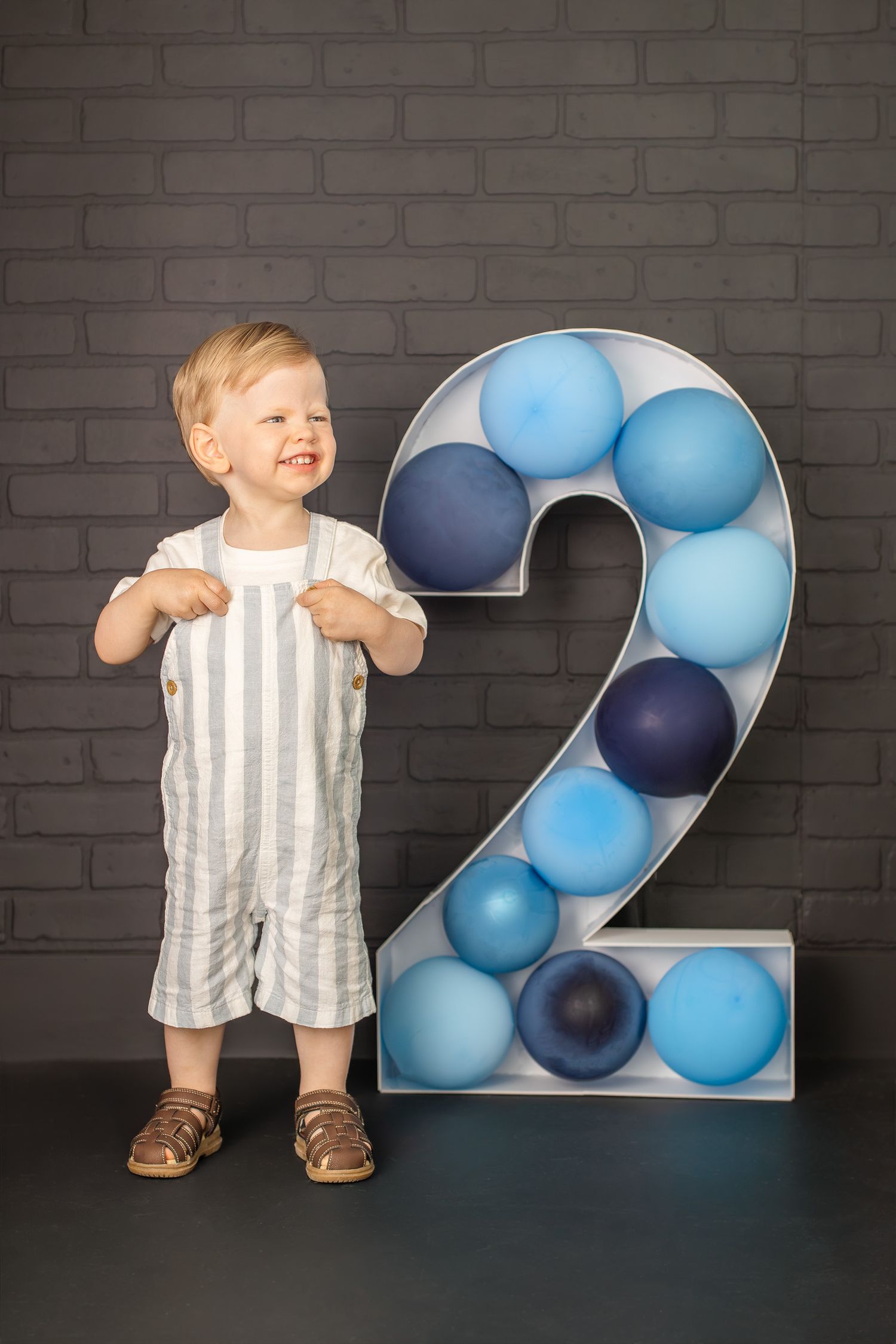 Toddler boy in striped overalls standing next to a large number 2 decorated with blue balloons against a brick wall.