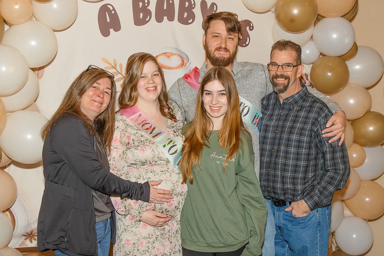 Family group photo at a baby shower with balloon arch backdrop, featuring pregnant woman wearing mom-to-be sash.