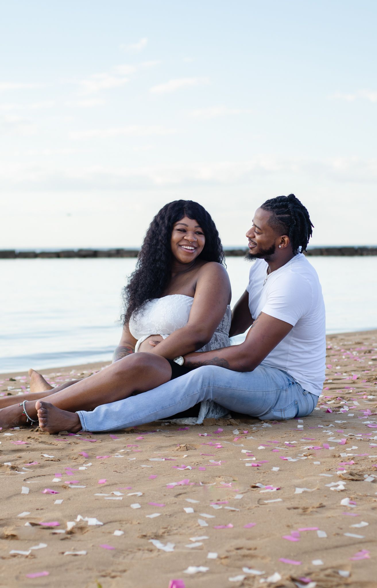 Couple sits on beach surrounded by scattered pink petals at sunset.