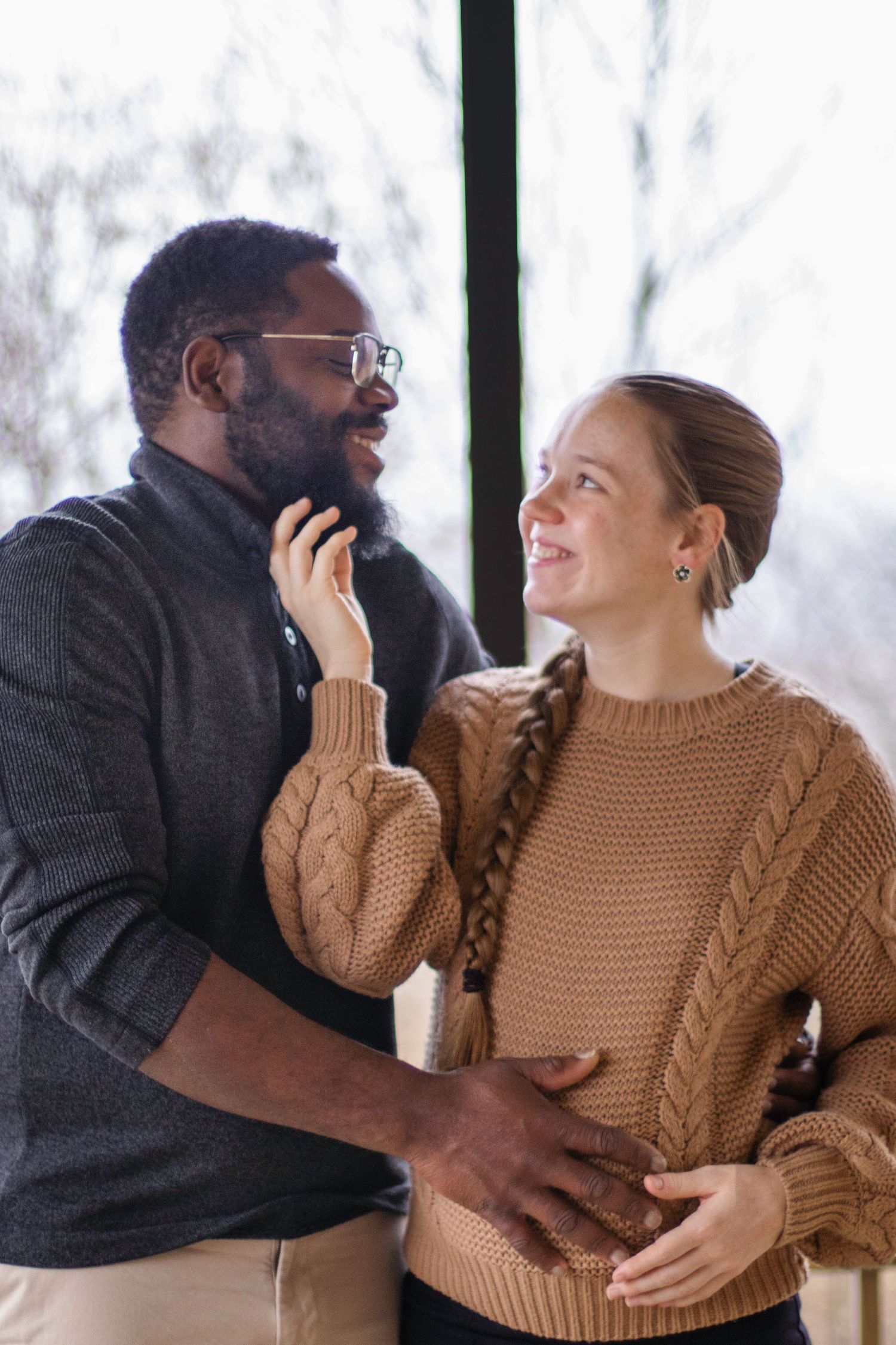 Expecting couple in cozy sweaters sharing an intimate moment by a window.
