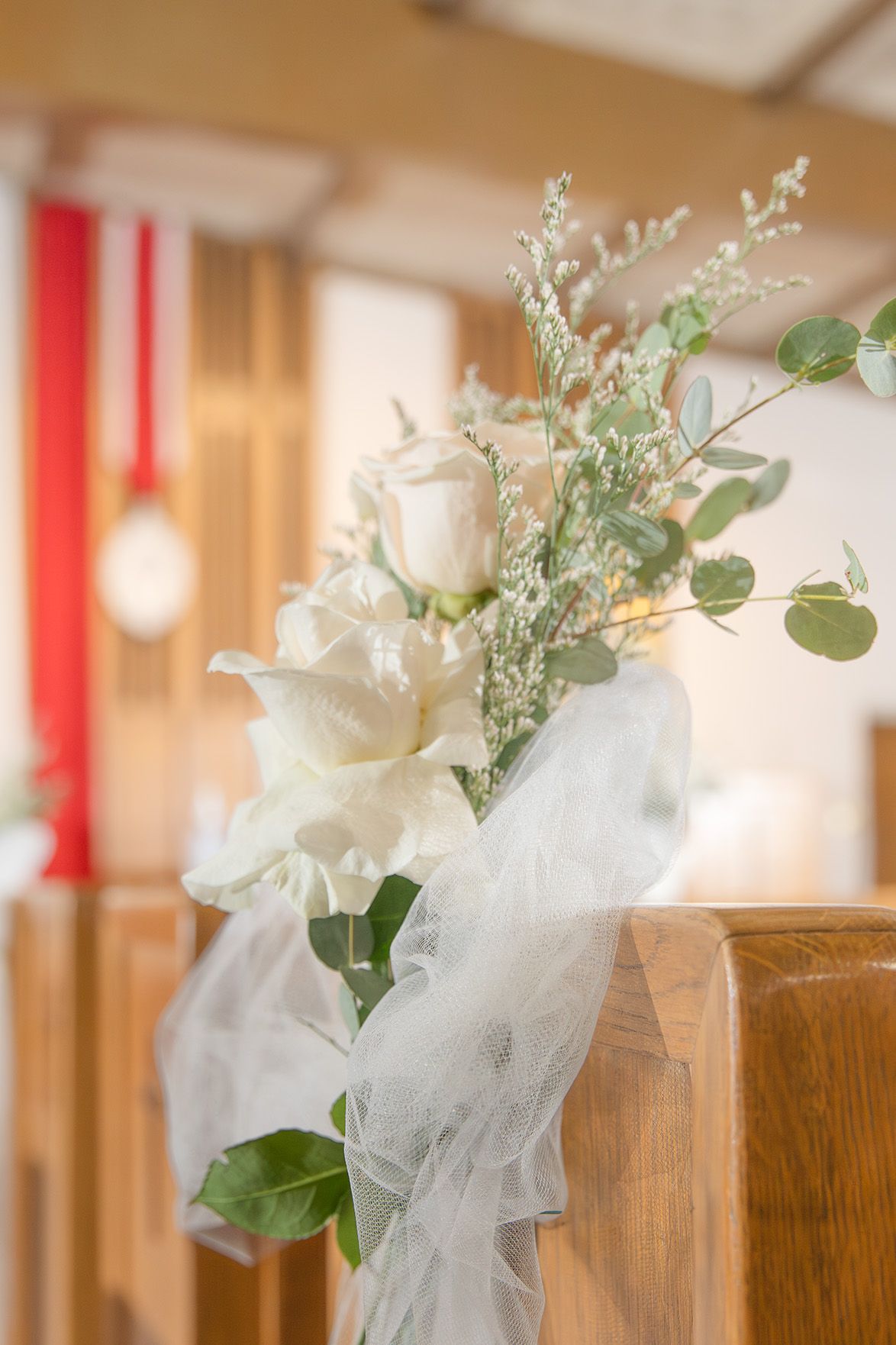 Elegant white roses and eucalyptus floral arrangement tied with tulle on church pew at wedding.