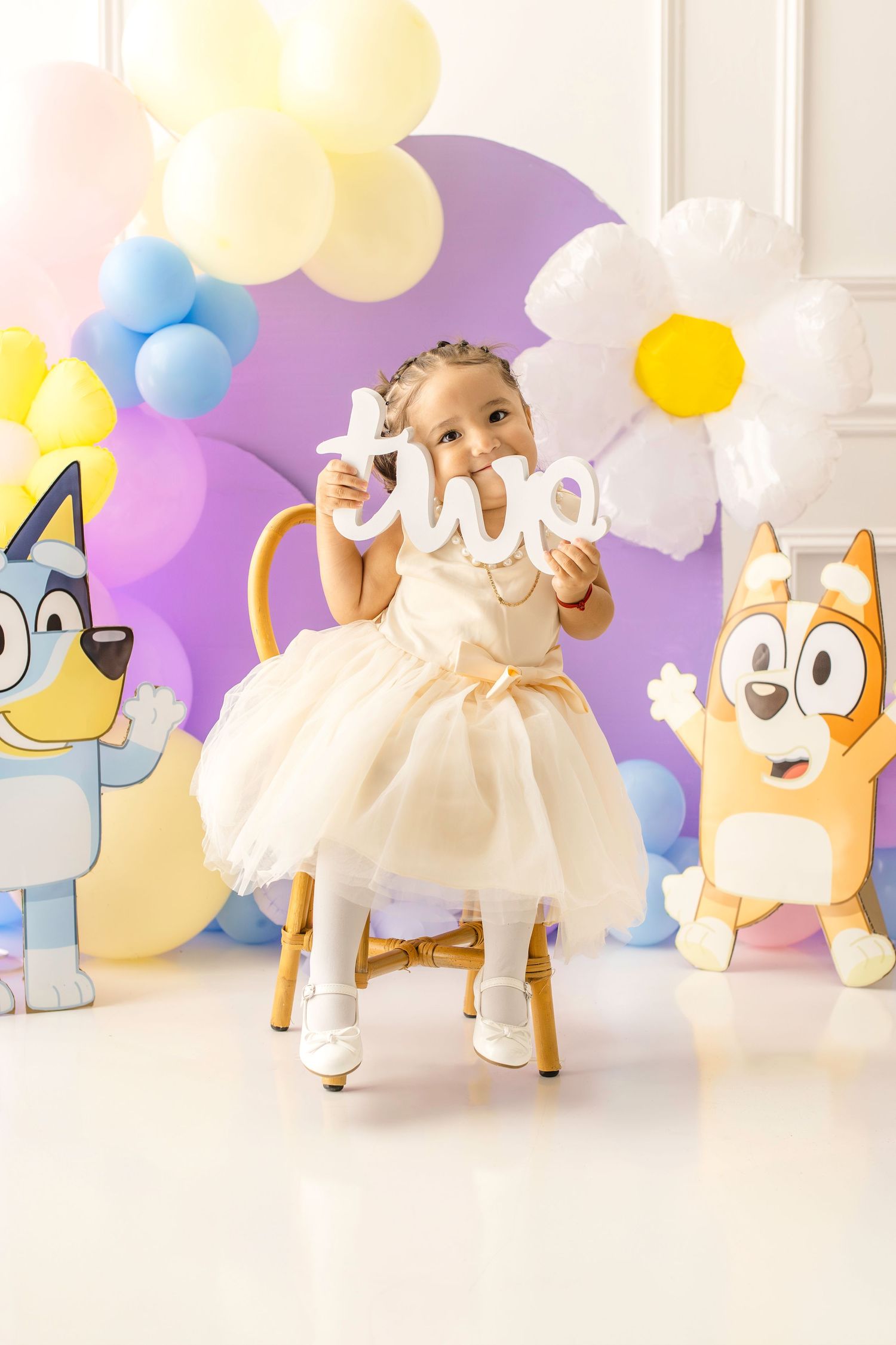 Toddler girl in white dress holds 'two' sign at Bluey-themed 2nd birthday party with colorful balloons.