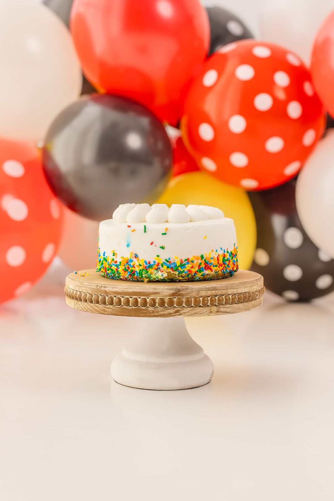 Small white birthday cake with colorful sprinkles on a wooden cake stand surrounded by red, black, and yellow balloons.