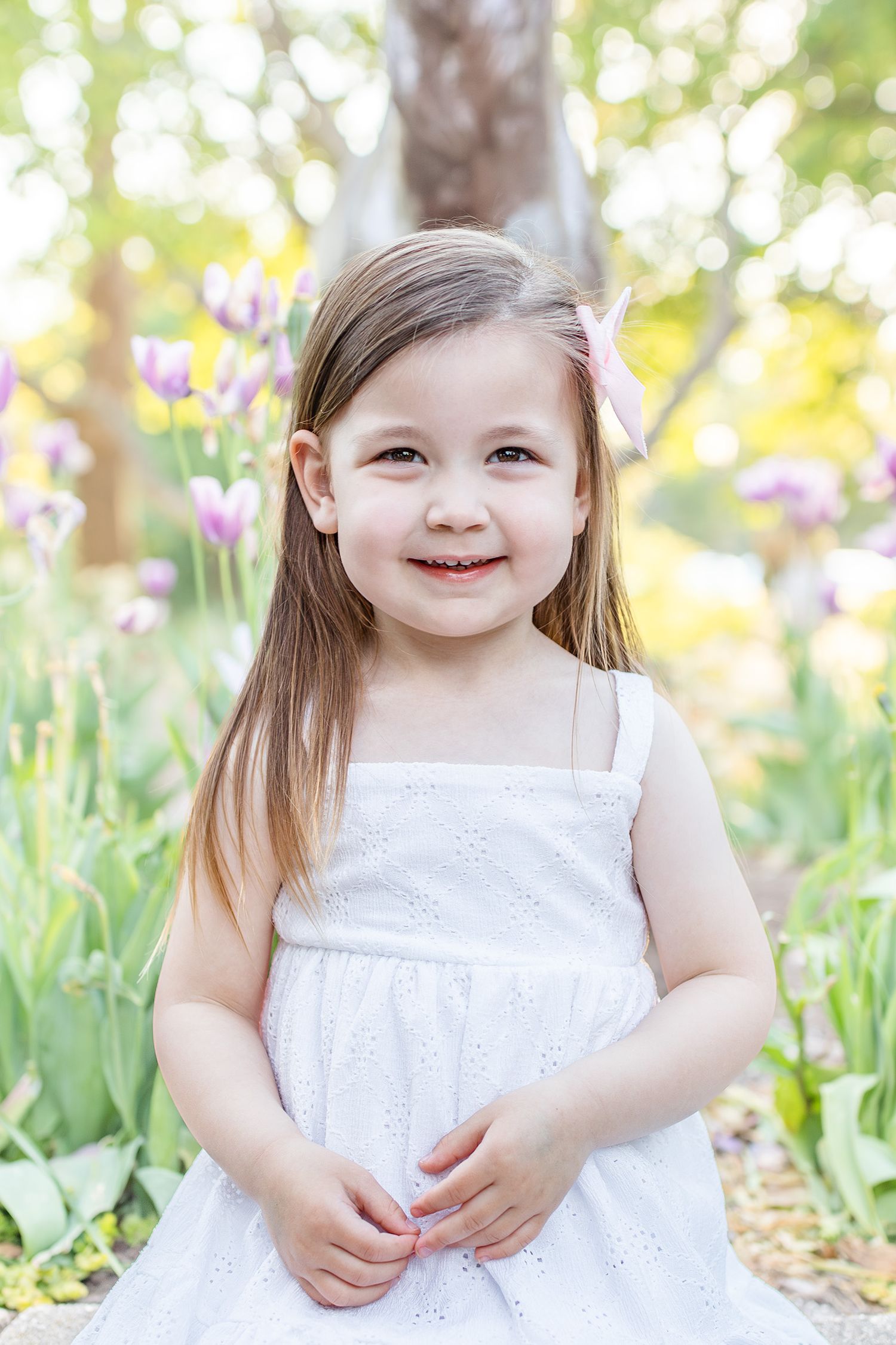 Smiling little girl in white dress standing among purple tulips in a sunny garden.