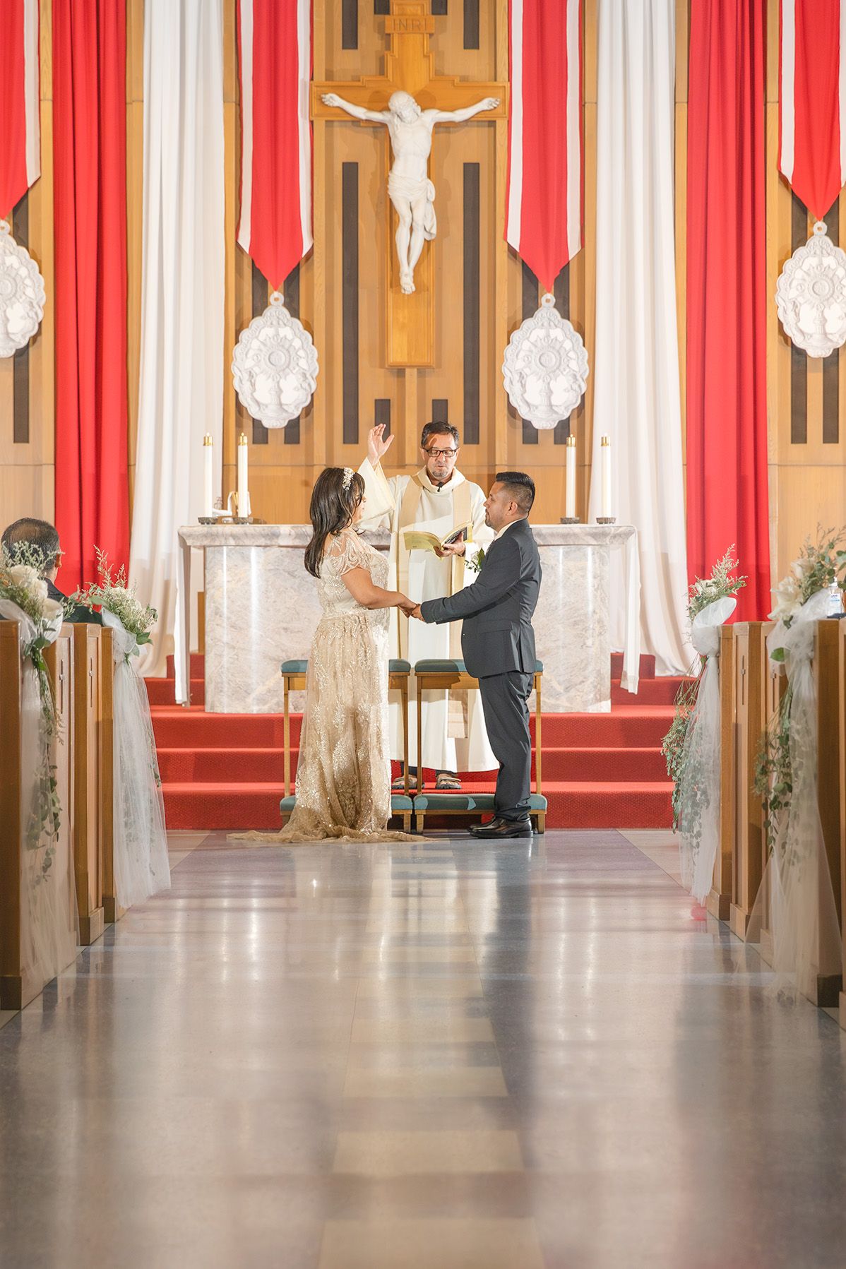 Couple exchanging vows at a Catholic church altar with red drapes and crucifix during a wedding ceremony.