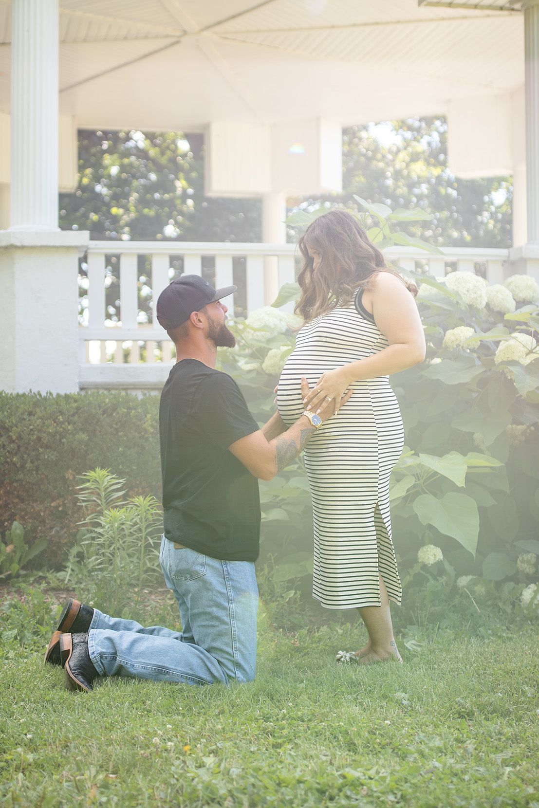 Man kneeling on grass touching pregnant womans belly in striped dress near white gazebo and hydrangeas.