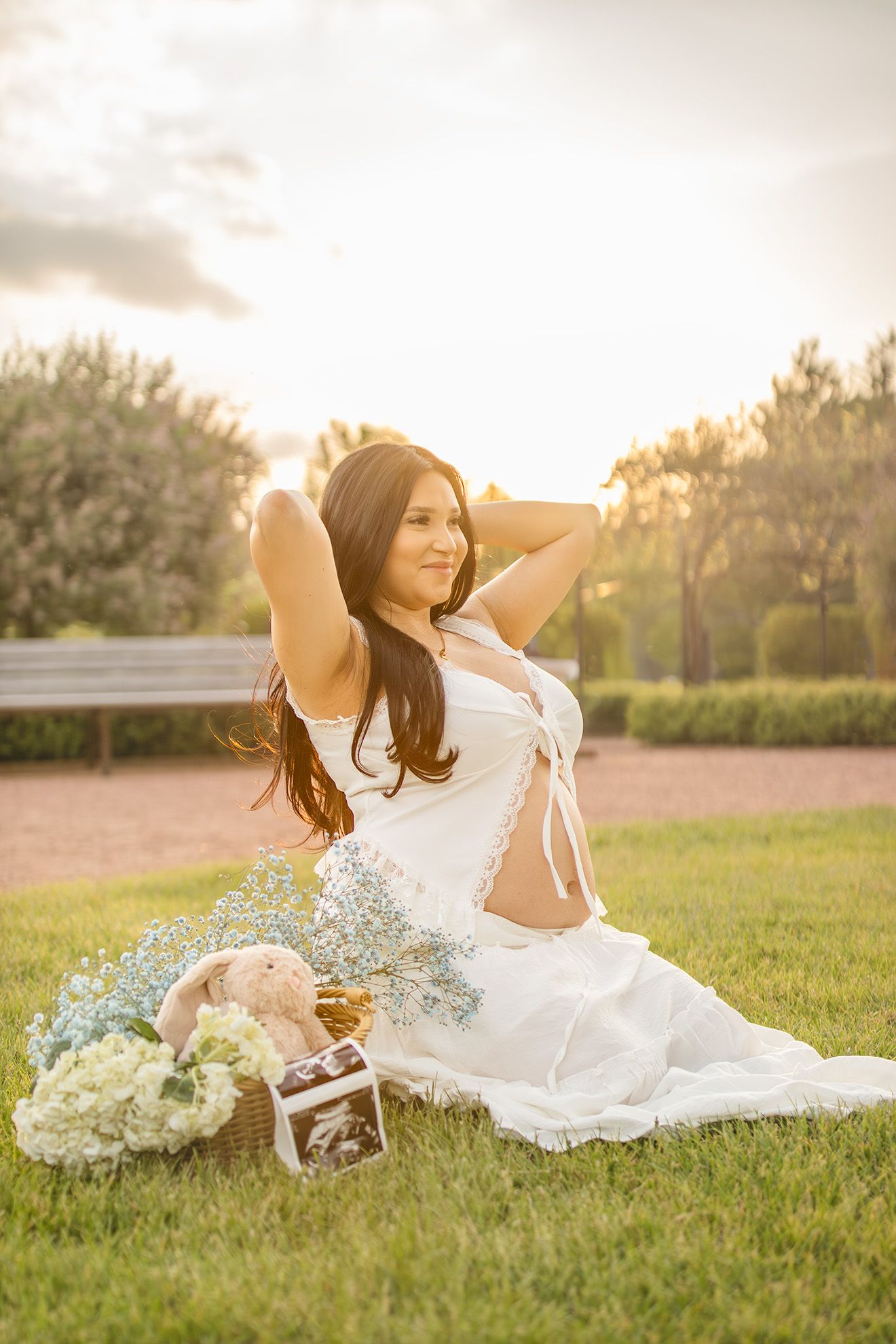 Pregnant woman in white dress sitting on grass with flowers and ultrasound photos at golden hour.