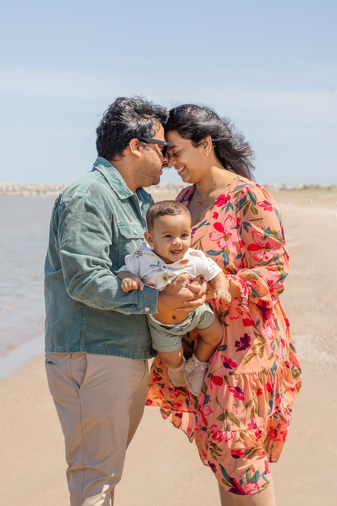 Happy family with baby enjoying a sunny beach day, parents touching foreheads while holding their smiling infant.