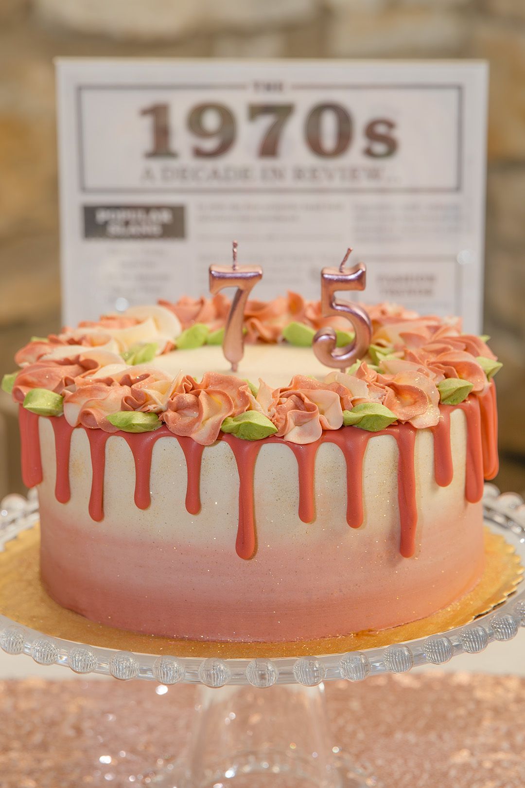 A 75th birthday cake with pink drip frosting and rose decorations, displayed in front of a 1970s decade in review sign.