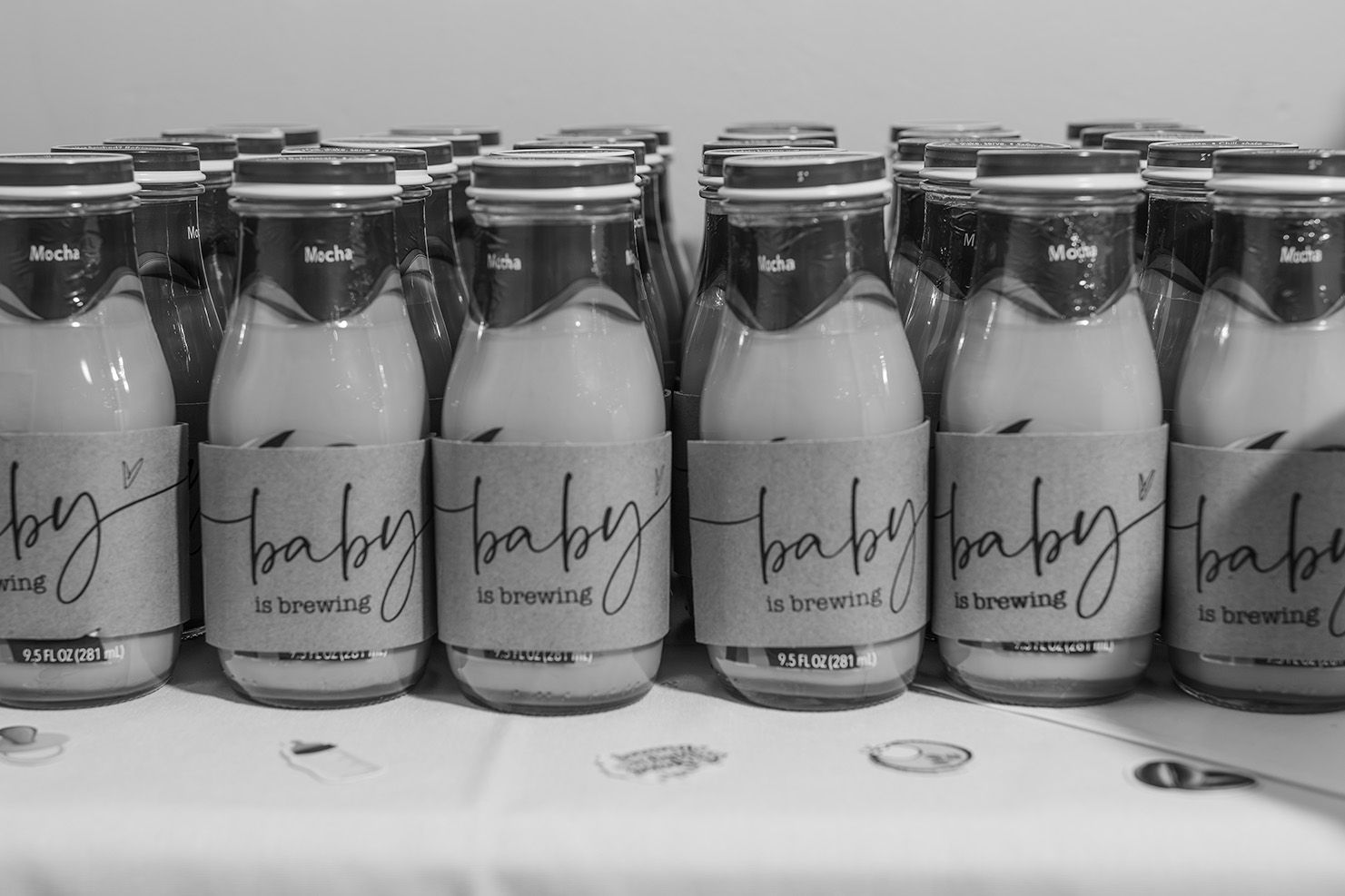 Black and white photo of baby shower milk bottles labeled baby is brewing arranged on a table.