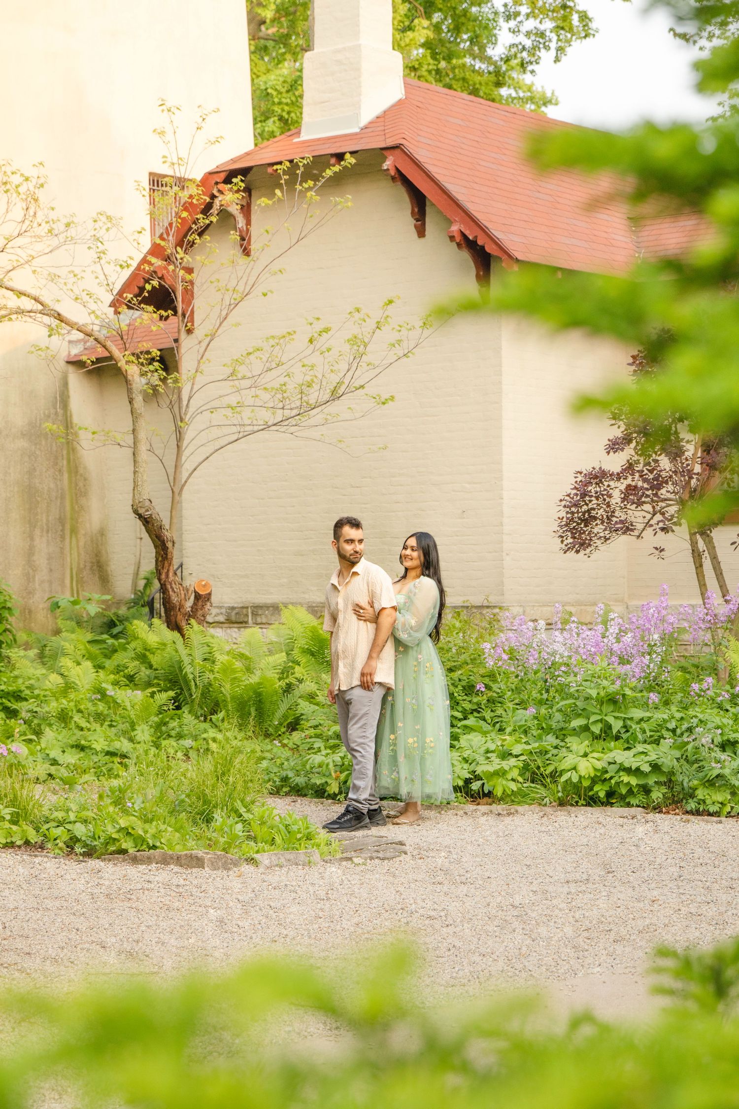 A couple stands together on a garden path beside a cream-colored building with a terracotta roof in springtime.