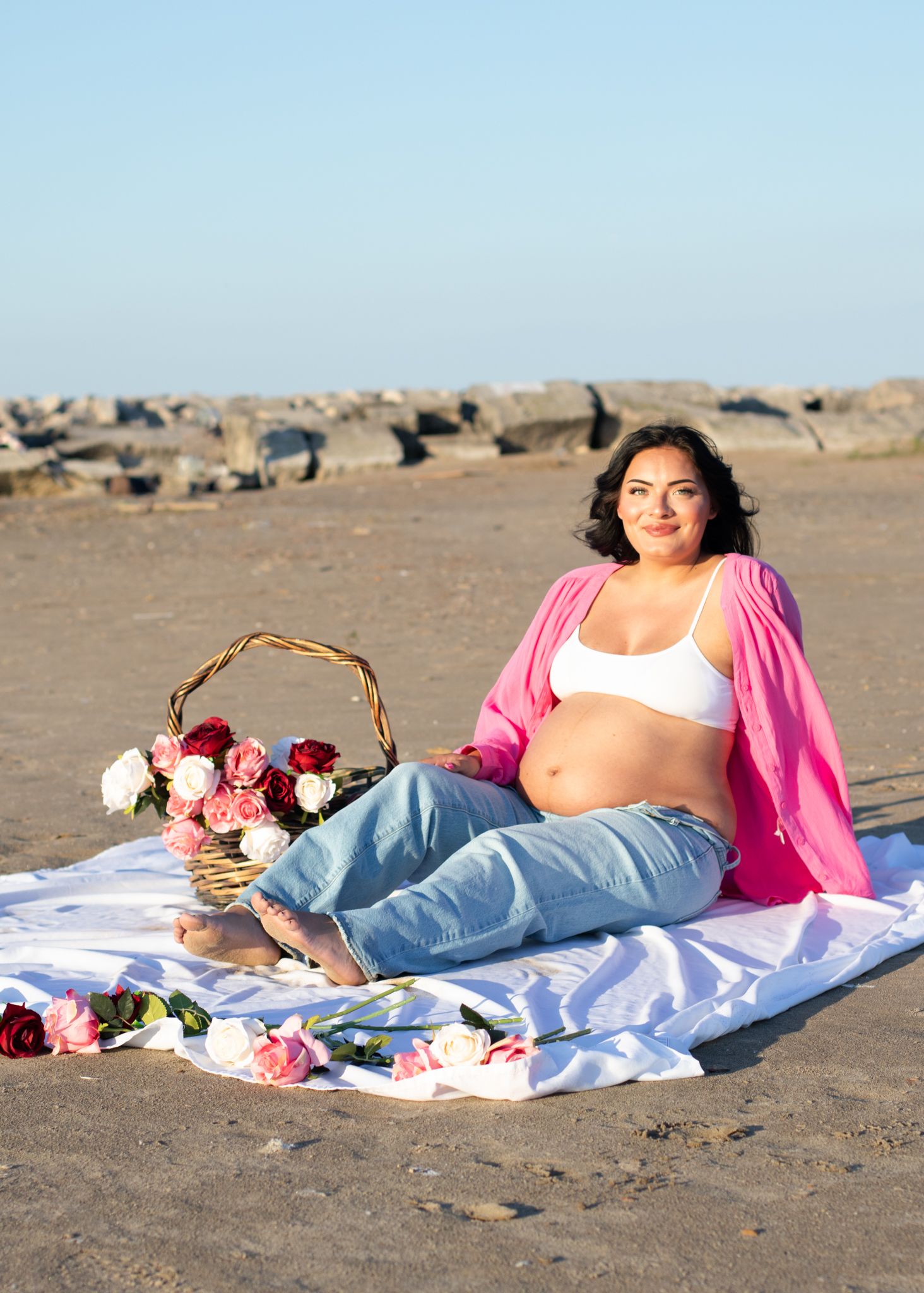 Maternity beach photo with pink cardigan, white blanket, and flower basket.