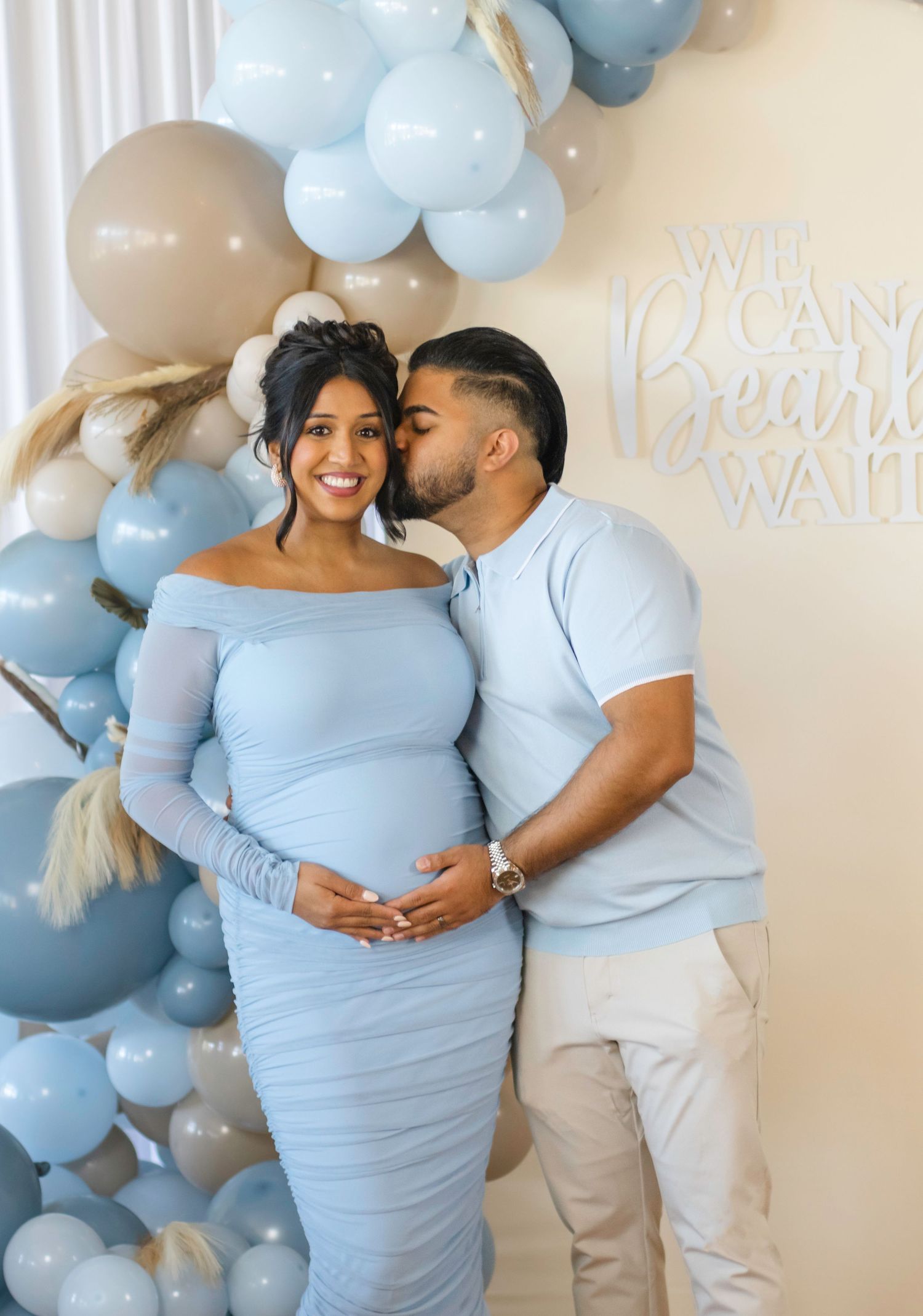 Maternity photo with balloon arch featuring couple in matching light blue outfits.