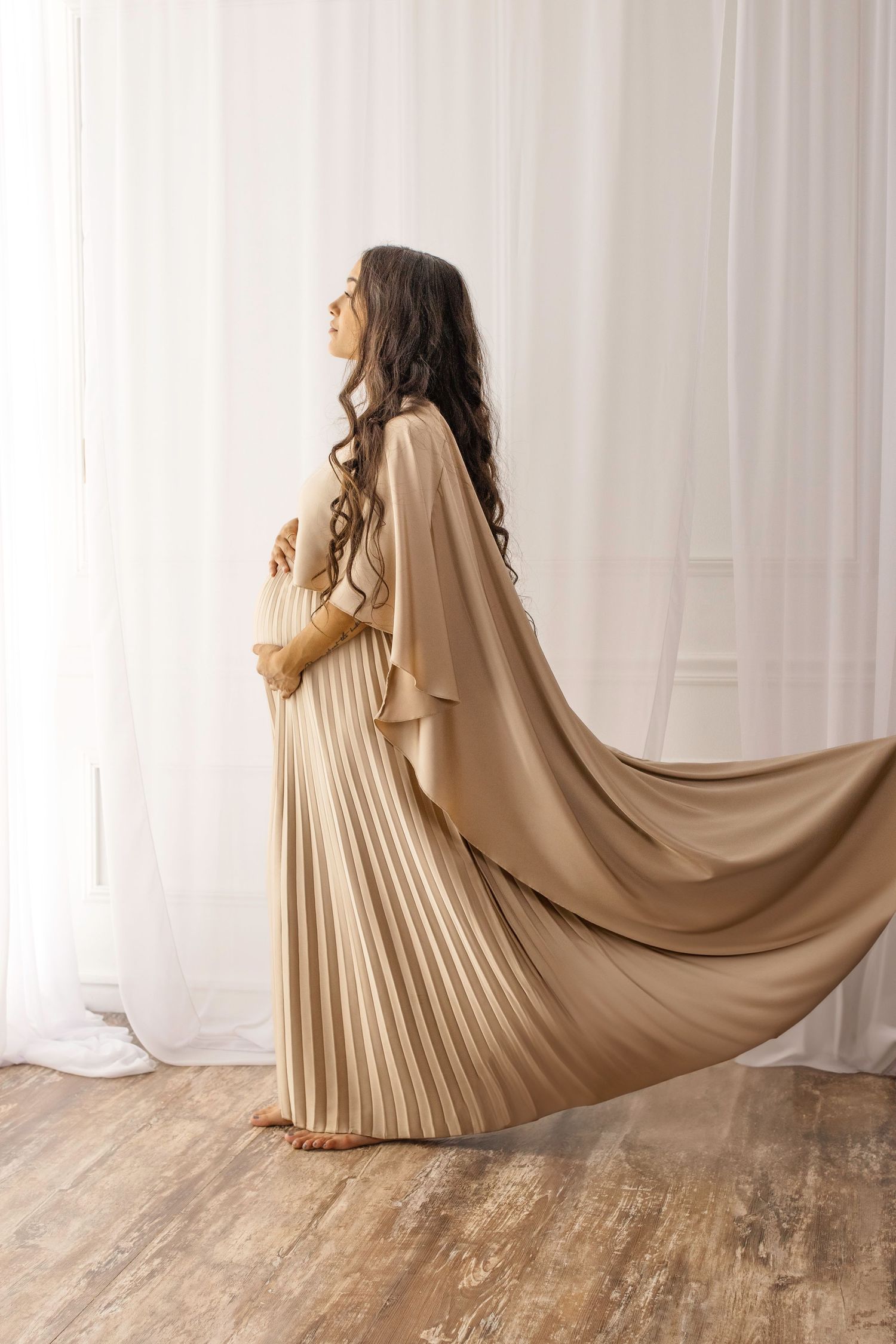 A person wearing a flowing beige pleated dress spins gracefully against a white curtain backdrop.