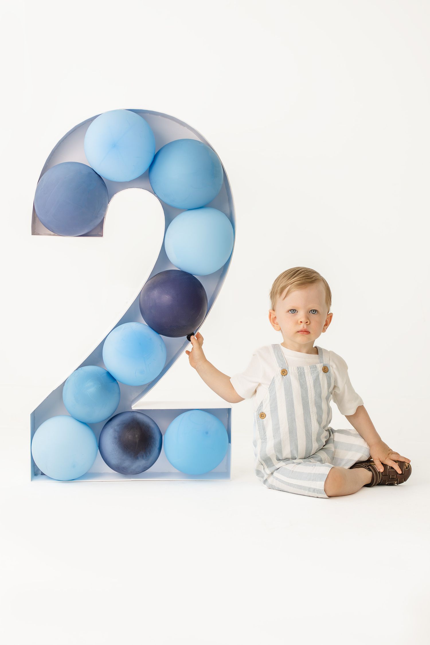 Toddler boy sitting next to a large number 2 filled with blue balloons for his second birthday photo.