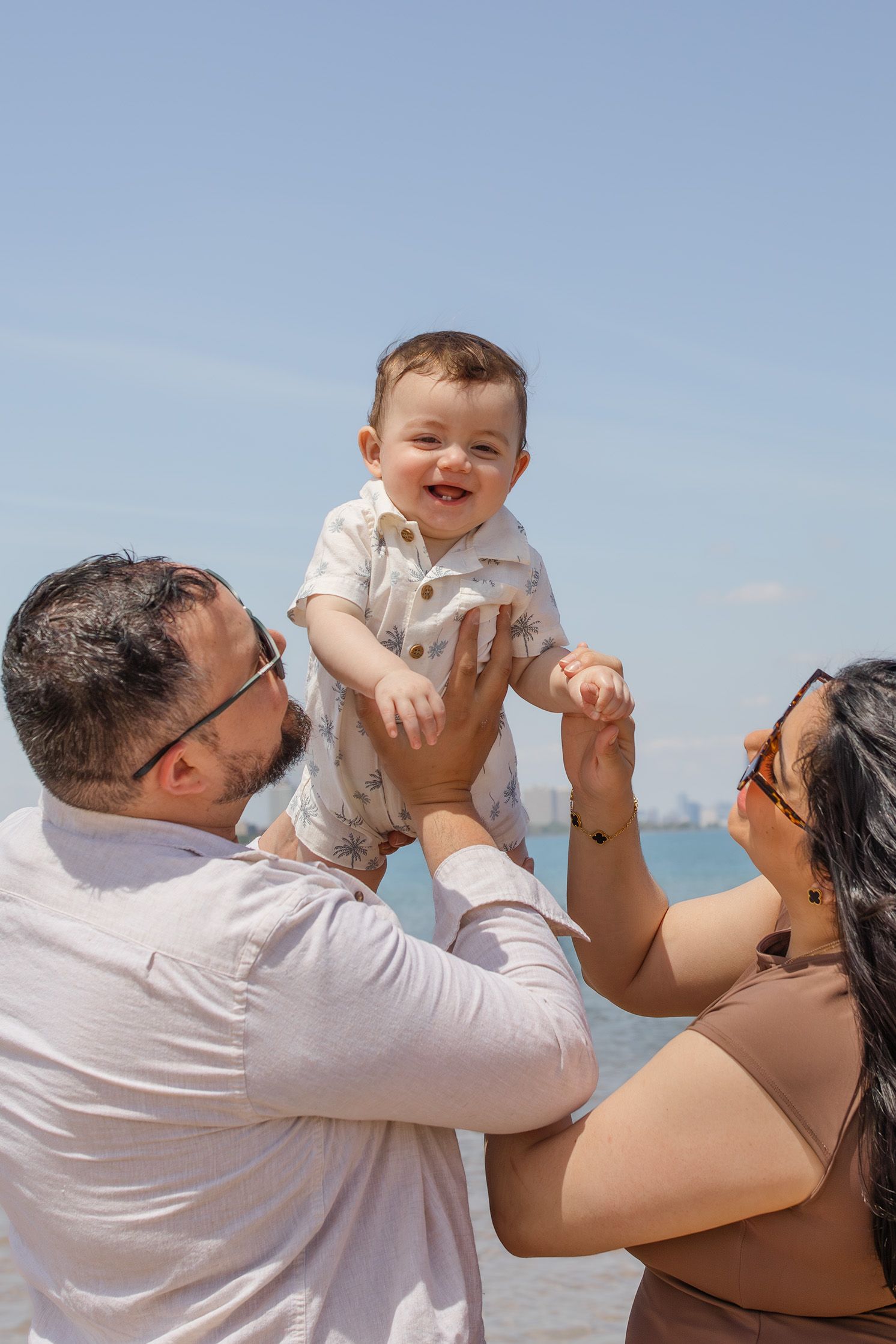 Happy baby laughing as parents lift him up at the beach on a sunny day.