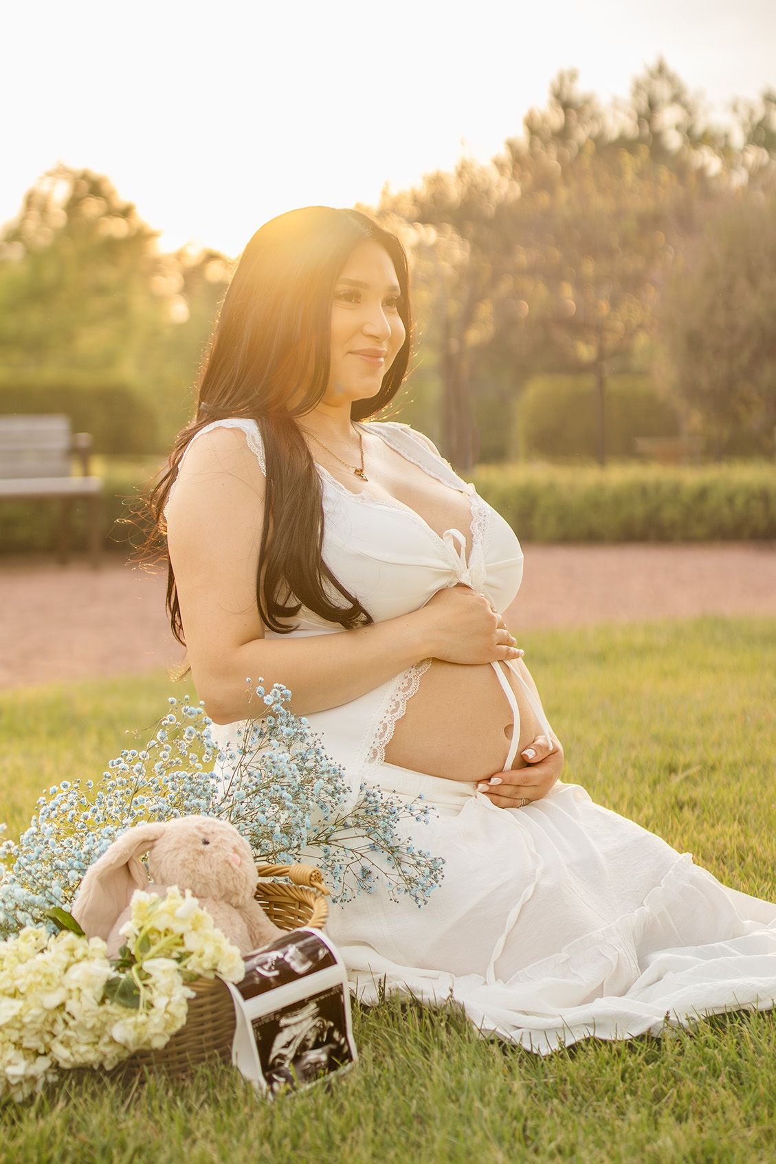 Pregnant woman in white dress sitting on grass at golden hour, cradling baby bump with flowers and ultrasound photos nearby.