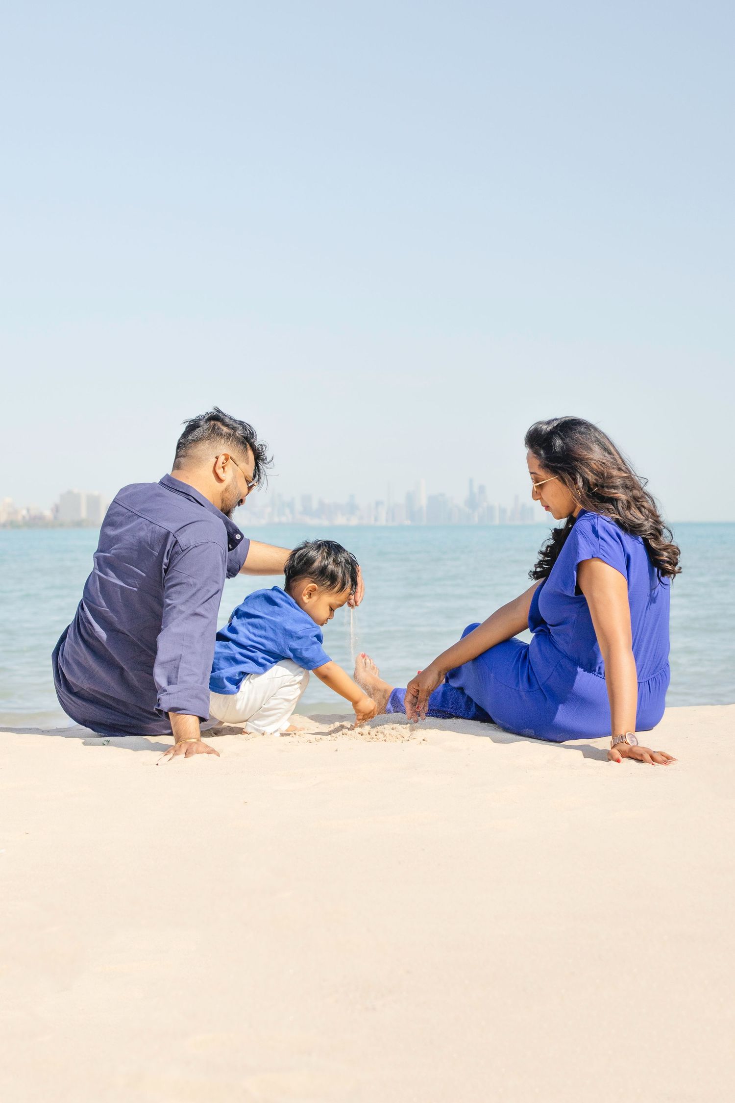 A family sits together on a sandy beach overlooking a city skyline across calm waters on a sunny day.