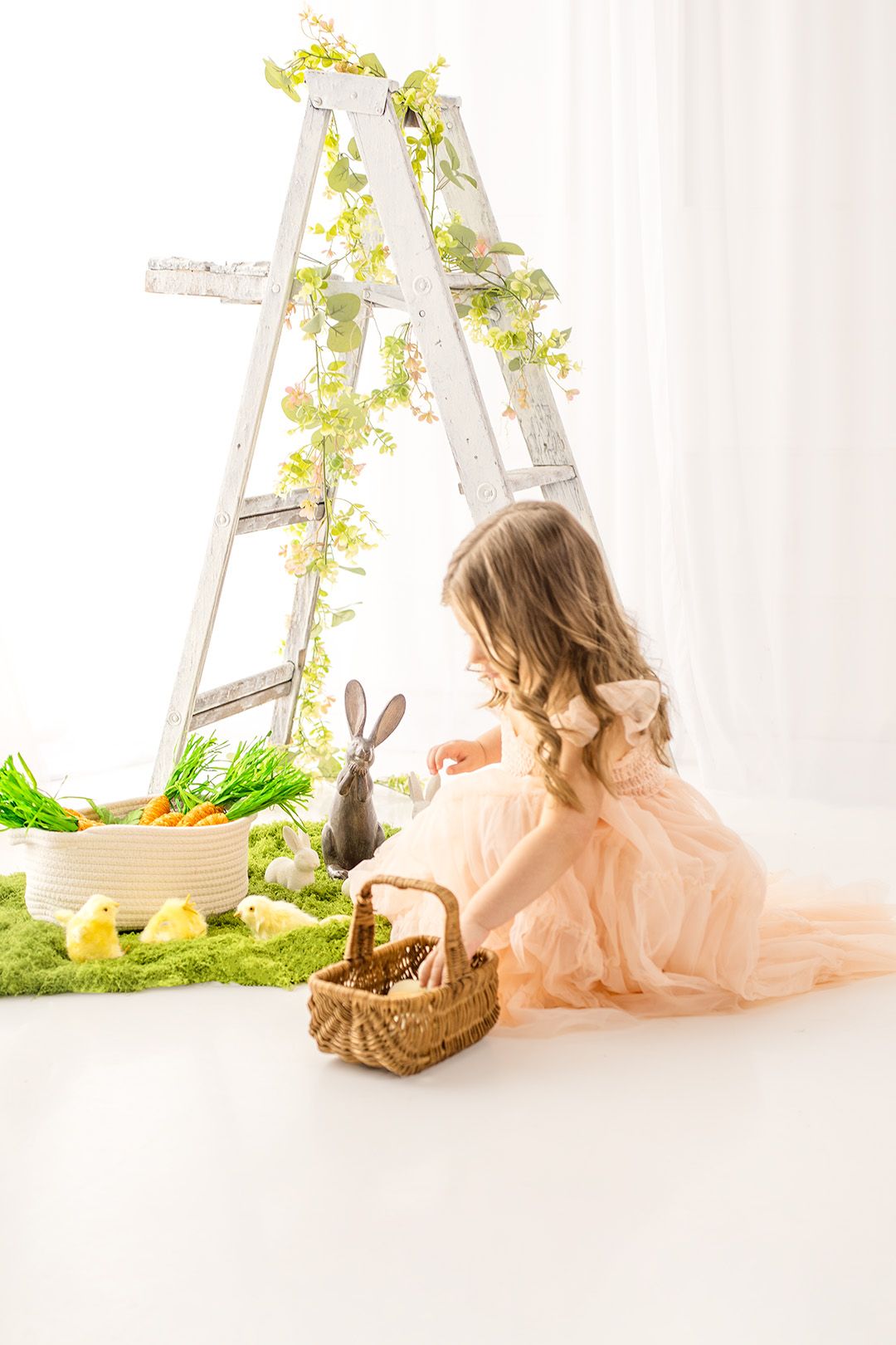A child in a peach dress sits near a rustic ladder with Easter decorations and spring greenery in a bright setting.