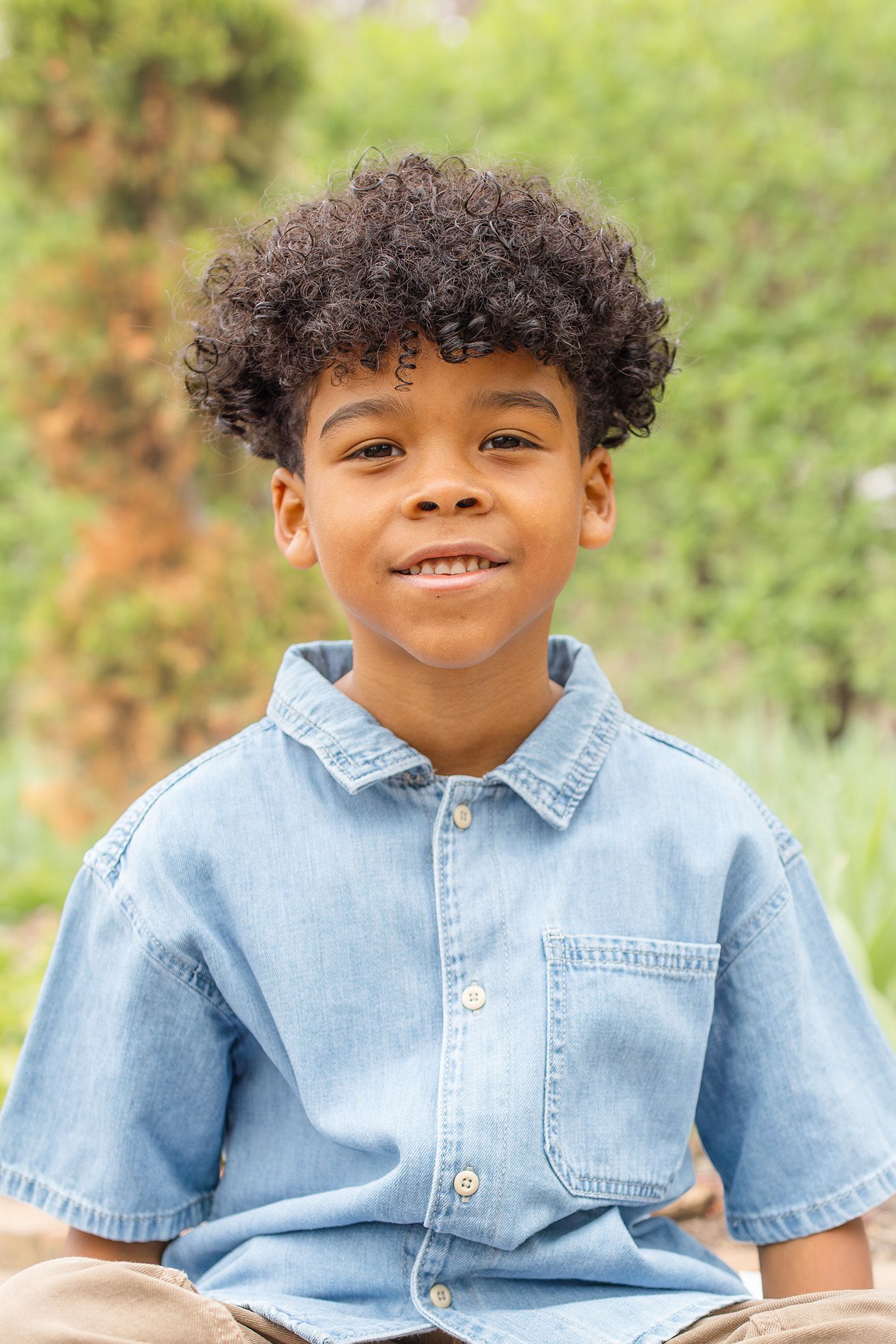 A young boy with curly hair smiles outdoors, wearing a light blue denim shirt with a green background.