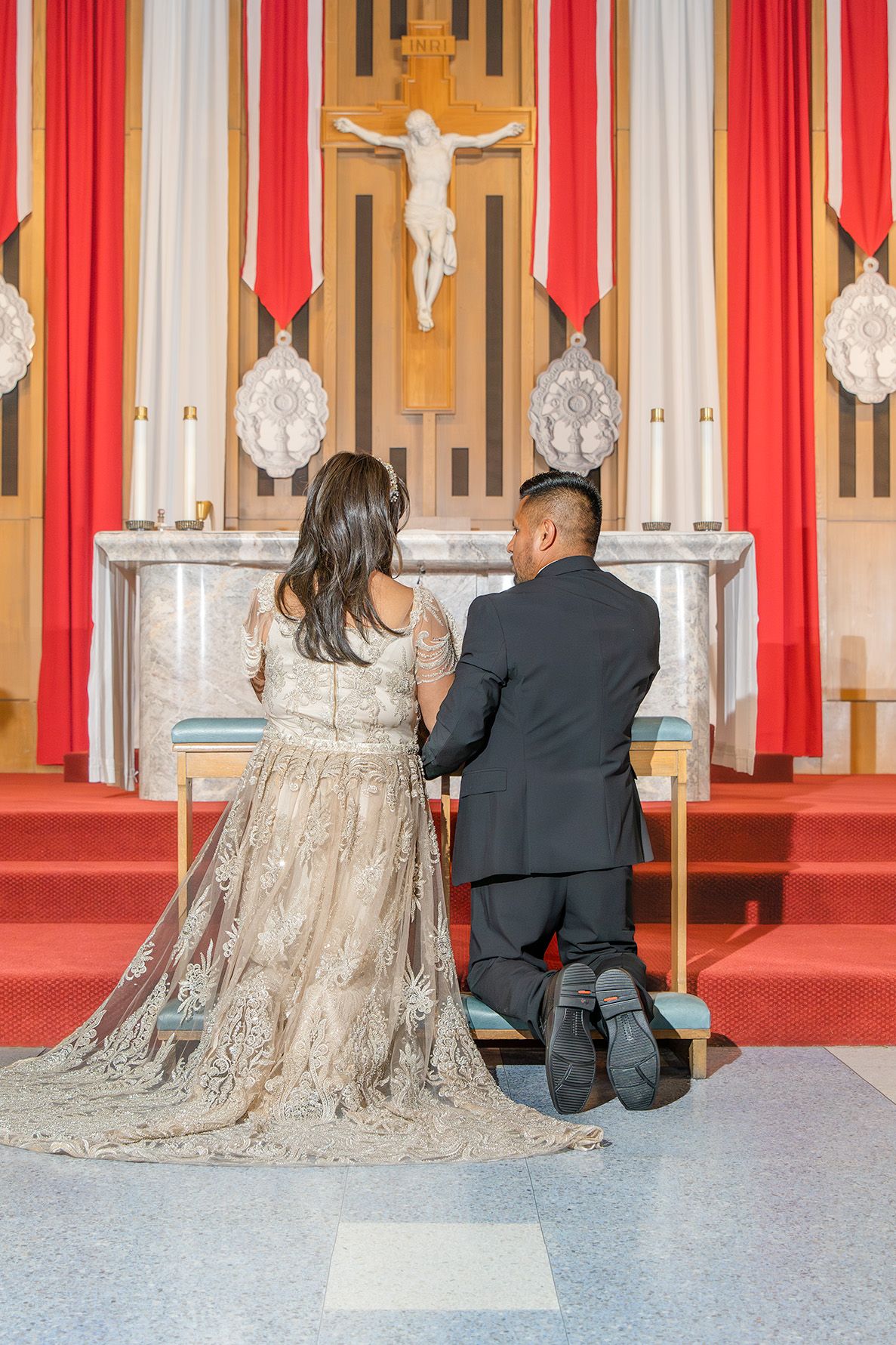 Couple kneeling in prayer before a church altar with crucifix, bride in lace gown and groom in black suit.