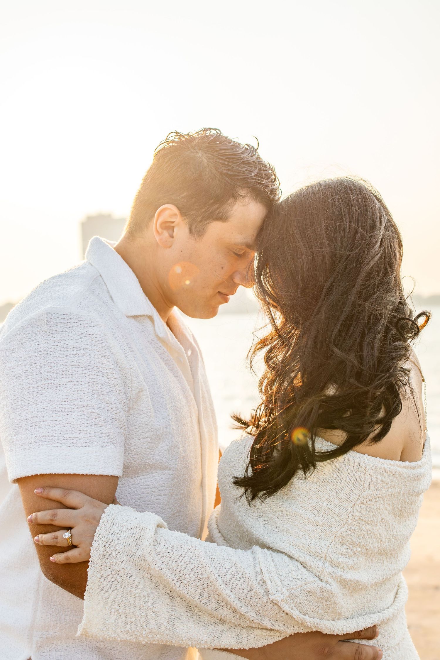 Couple in white outfits embracing on a beach at golden sunset, sharing a romantic moment with warm backlight.