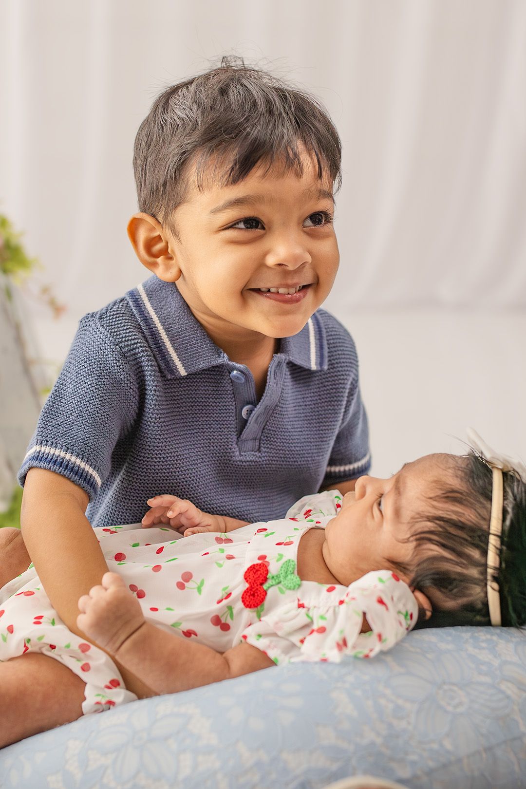 Smiling toddler boy in blue polo shirt sitting with newborn baby sibling in floral onesie on a soft surface.