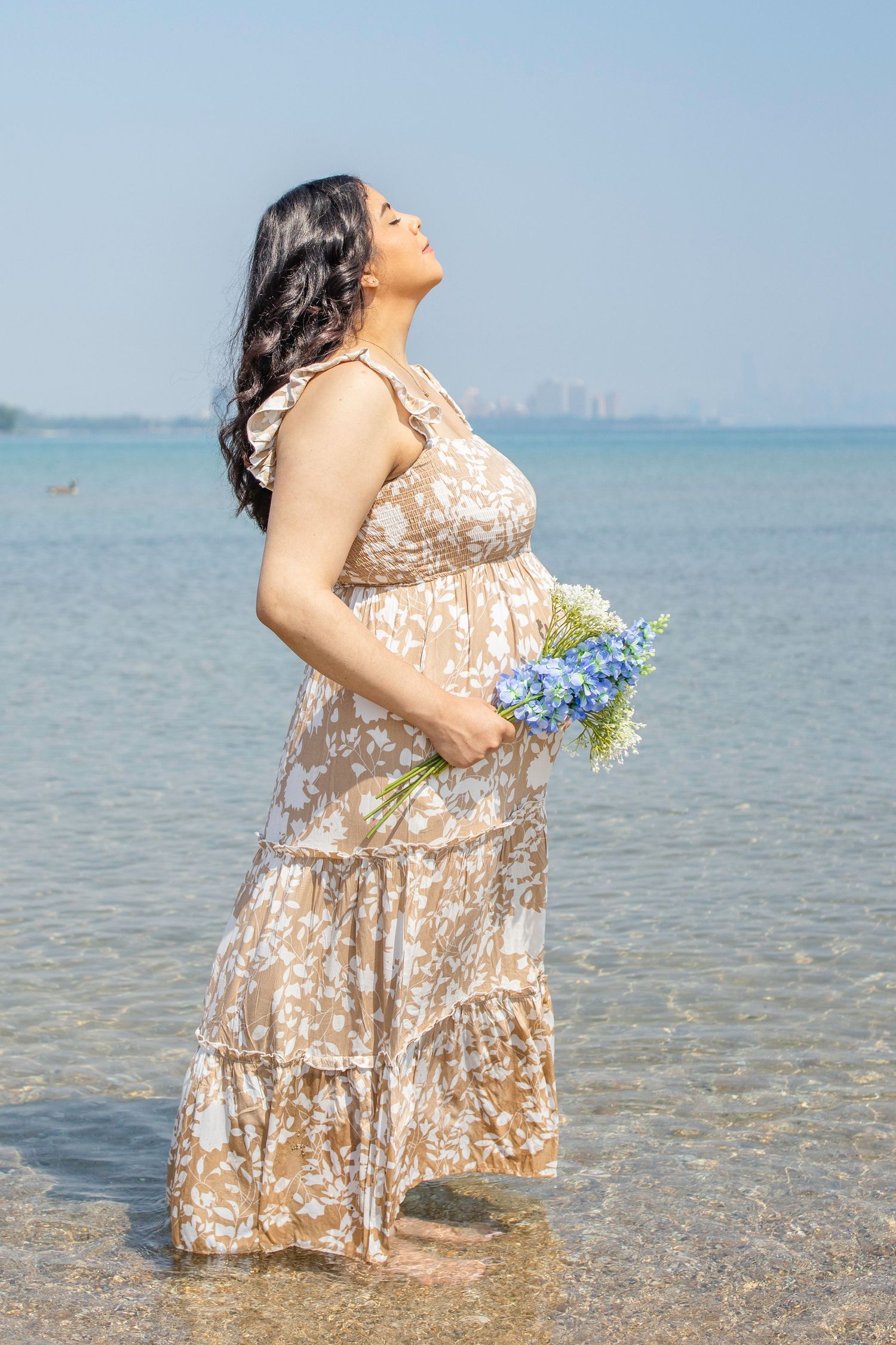 Pregnant woman in floral maxi dress holds blue flowers while standing in shallow ocean water on sunny day.