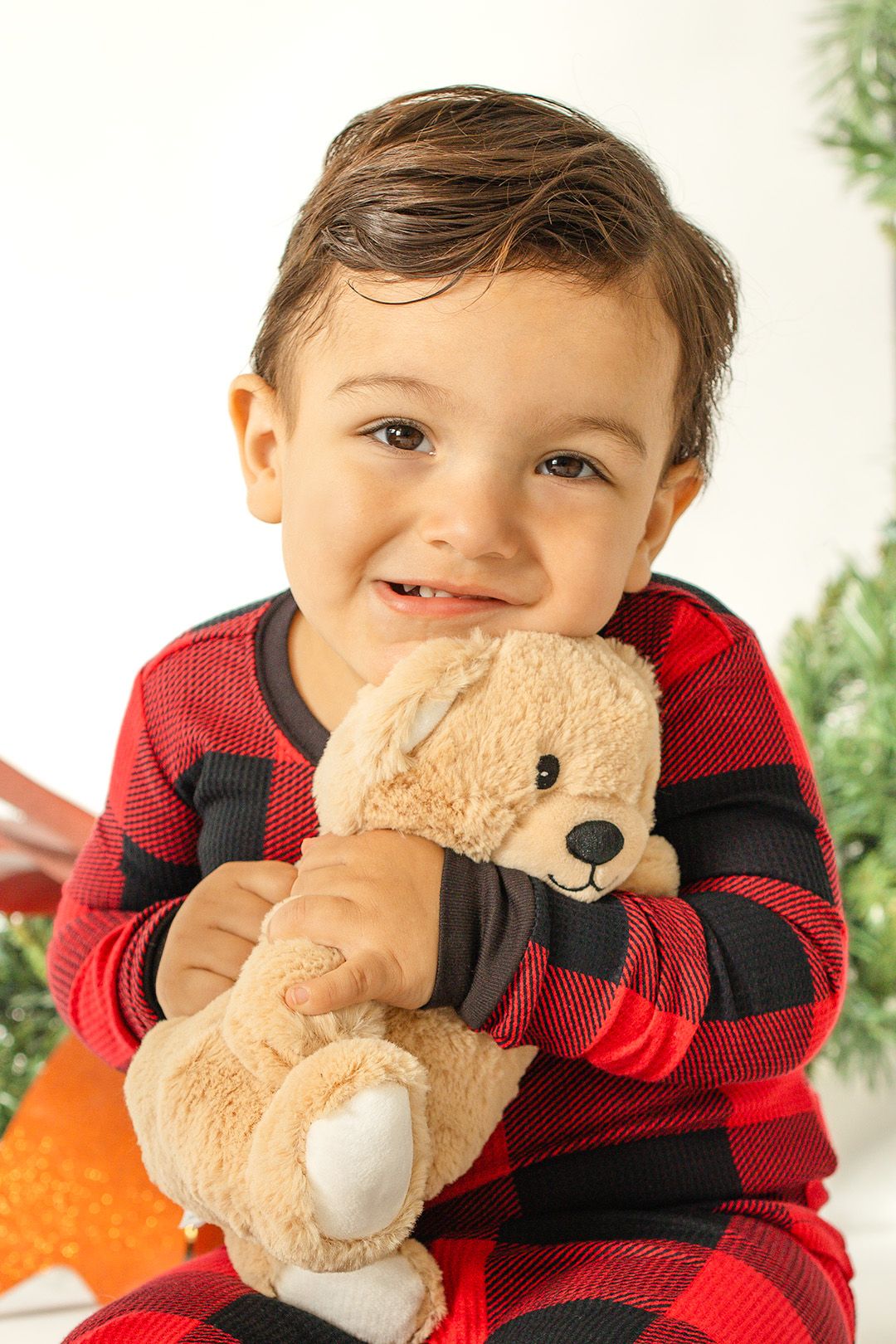 A child in a red and black buffalo plaid shirt holds and plays peek-a-boo with a soft tan teddy bear.