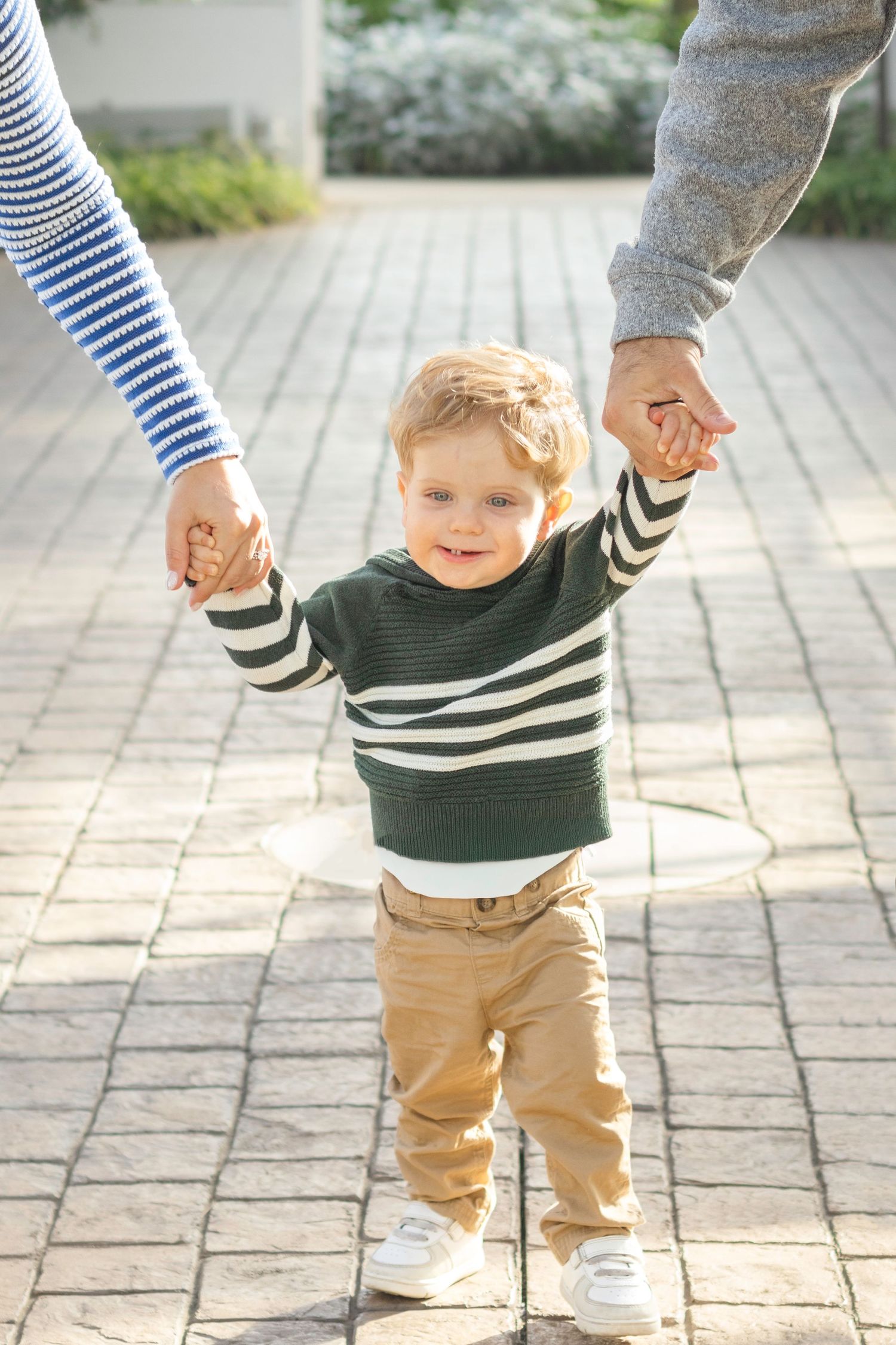 A toddler in a striped sweater holds hands with parents while walking on a stone path.