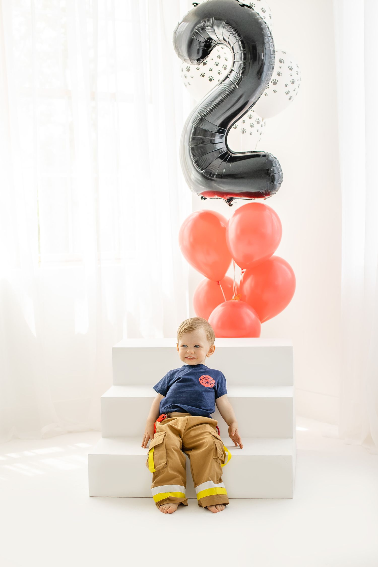 Toddler boy in firefighter costume sitting on white steps with red balloons and number 2 balloon.
