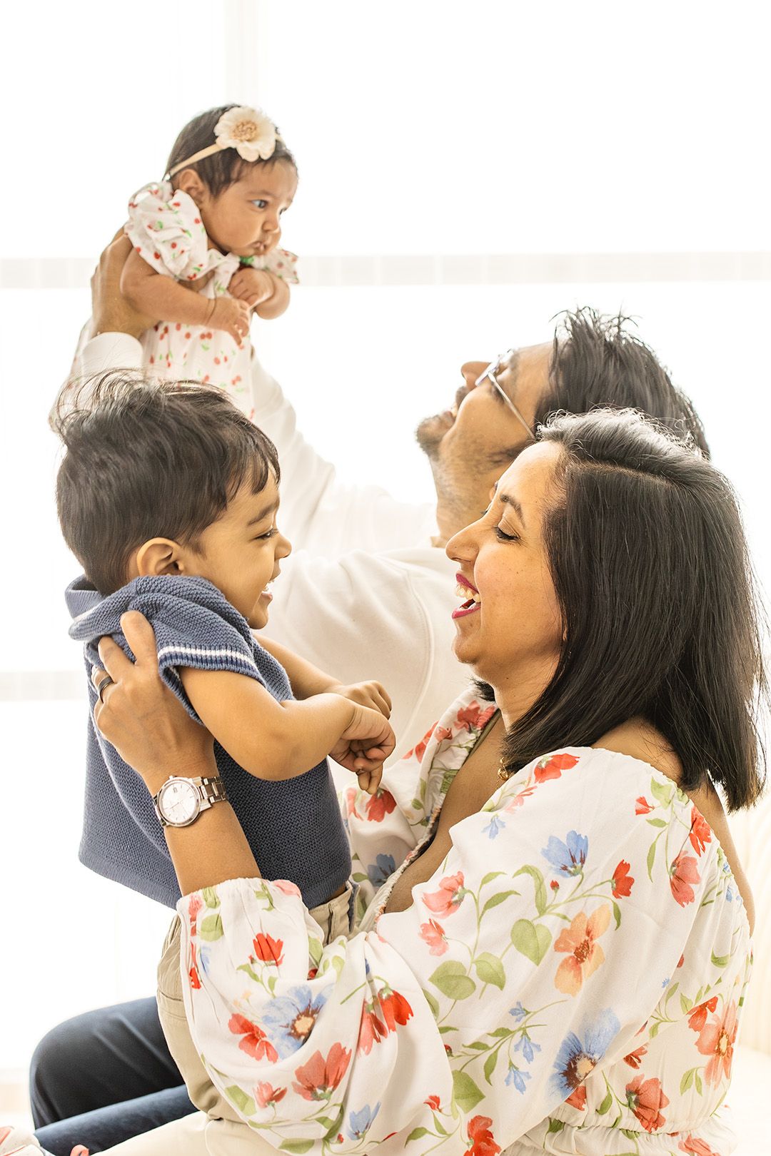 Happy South Asian family with parents holding two young children, a toddler boy and baby girl, in a bright room.
