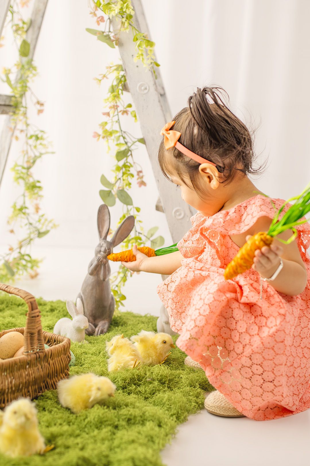 A young girl in a pink dress feeds toy carrots to rabbit figurines surrounded by baby chicks on green moss in an Easter scene.