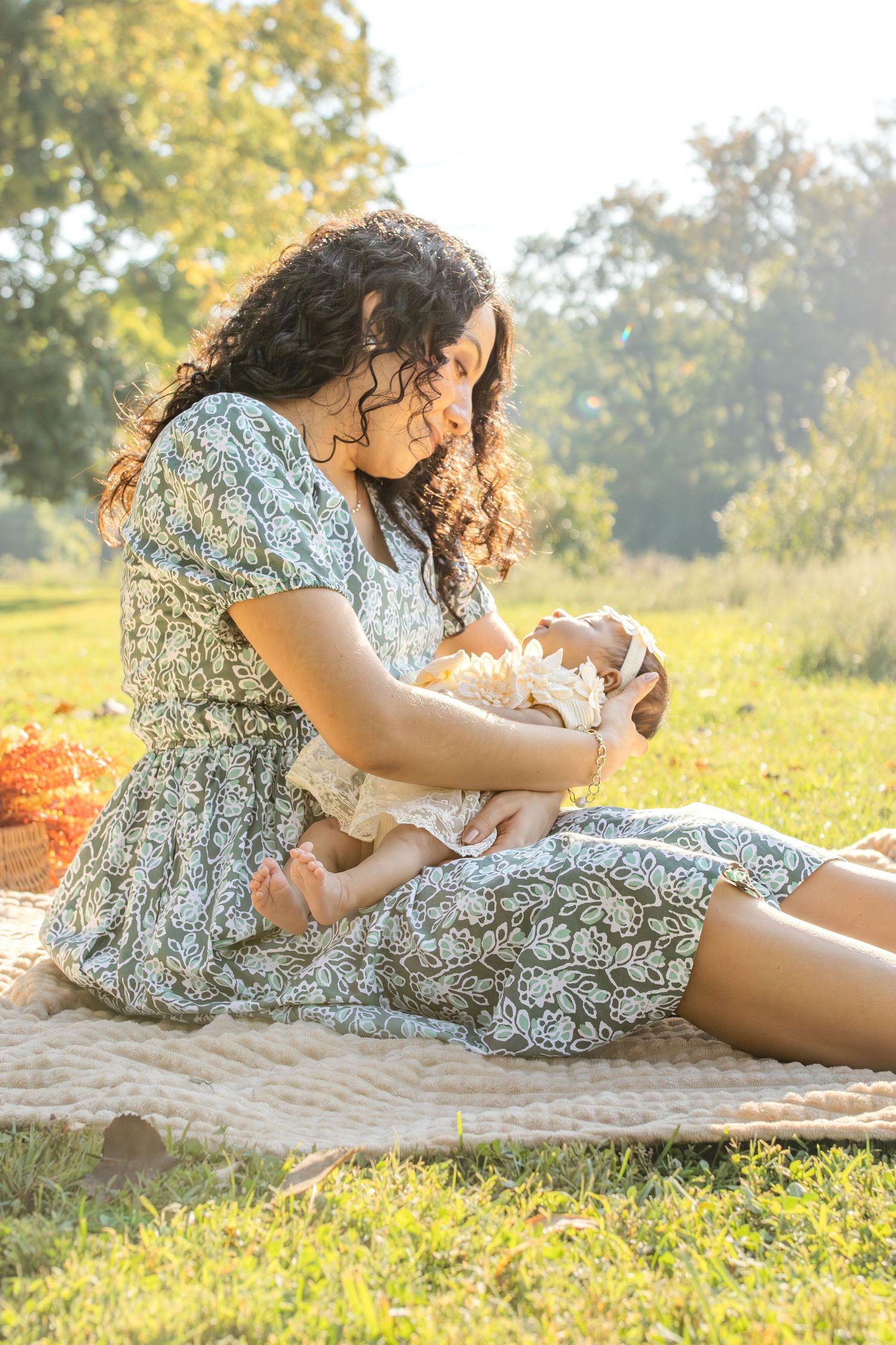 Mother reclines on blanket outdoors while tenderly holding newborn baby in afternoon sunlight.