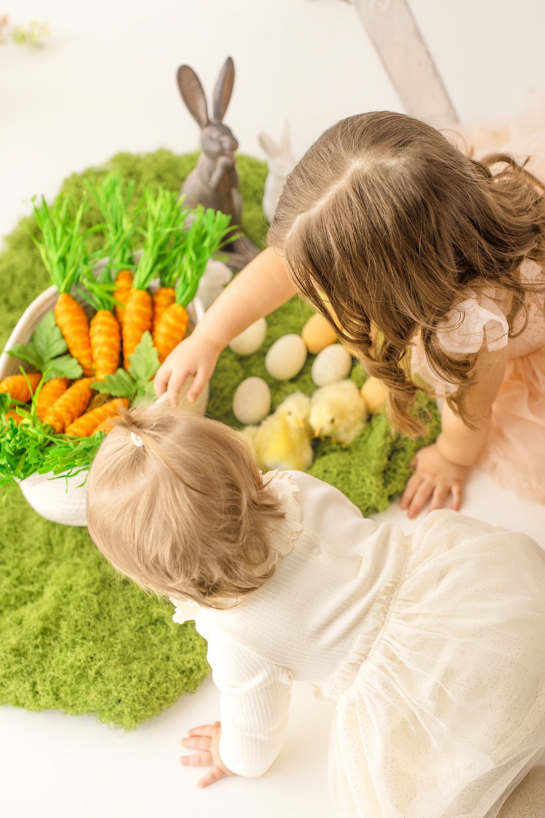 A child lies on a green carpet reaching toward decorative carrots and bunny toys in an Easter-themed setting.