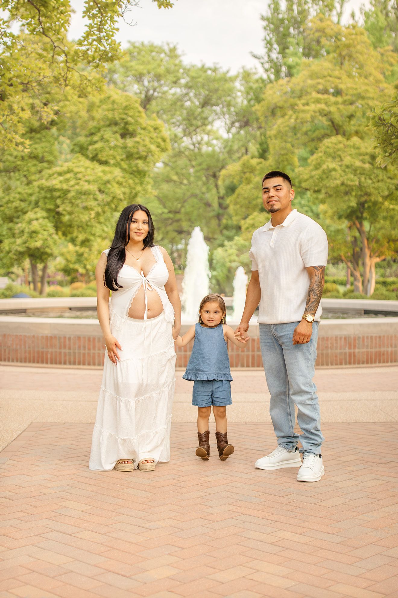 Pregnant woman in white dress with toddler and man in blue jeans holding hands by park fountain.