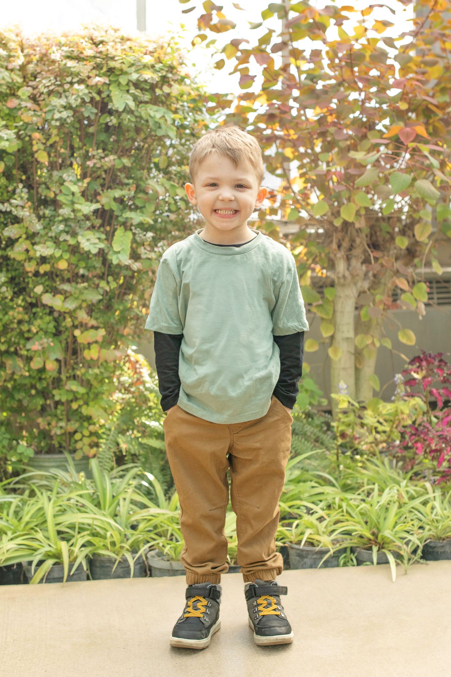 Young child in layered green shirt and khaki pants stands smiling in a garden setting.