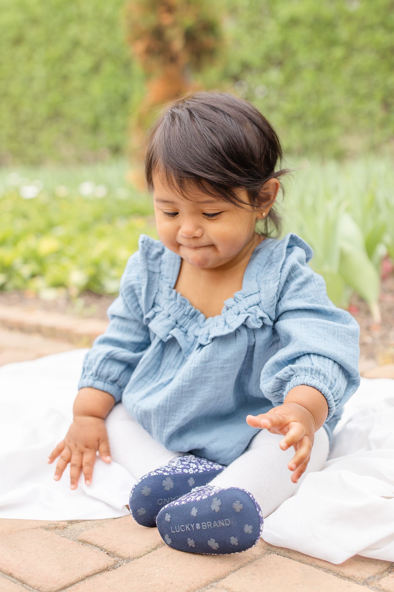 Toddler girl in blue ruffled top and Lucky Brand shoes sits outdoors on white blanket, smiling.